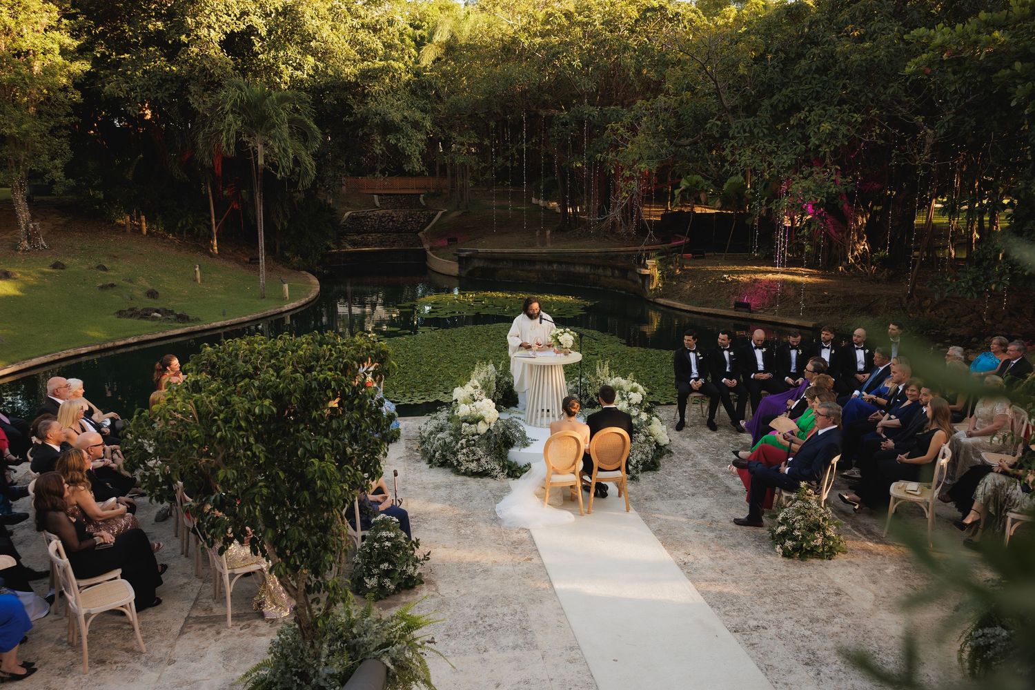 Wedding guests gather outdoors for an intimate backyard ceremony at twilight in a wooded garden setting.