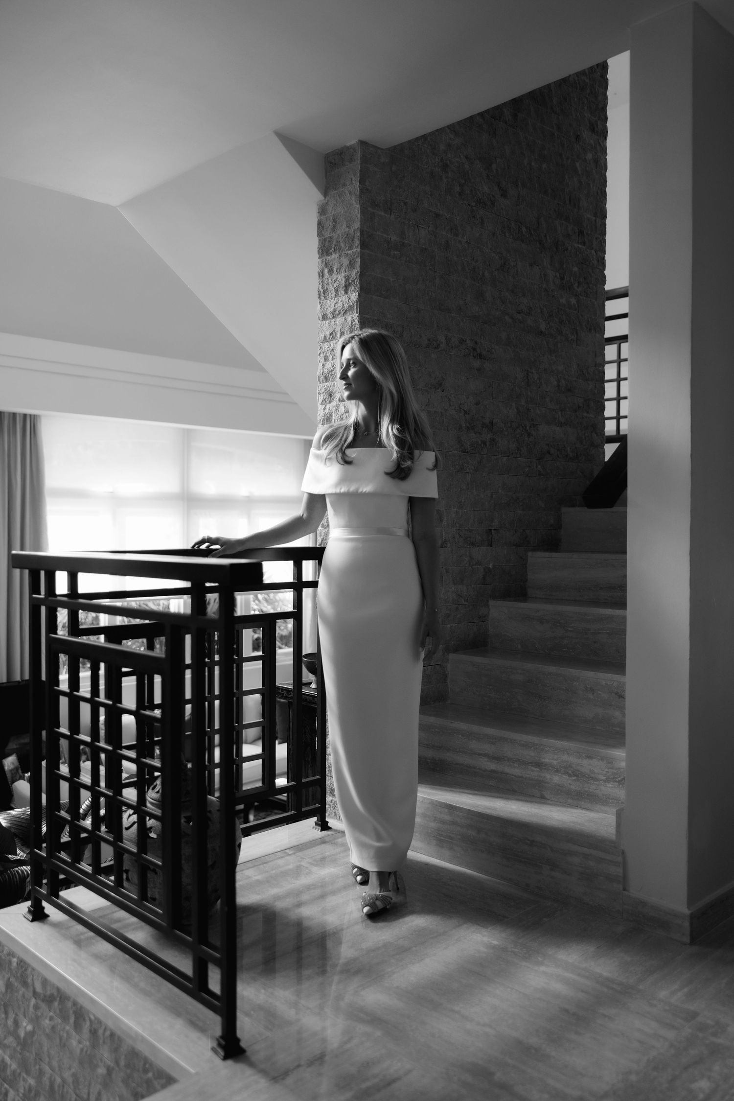 Black and white photo of a person in a fitted white dress standing near a modern staircase and railing in an interior space.