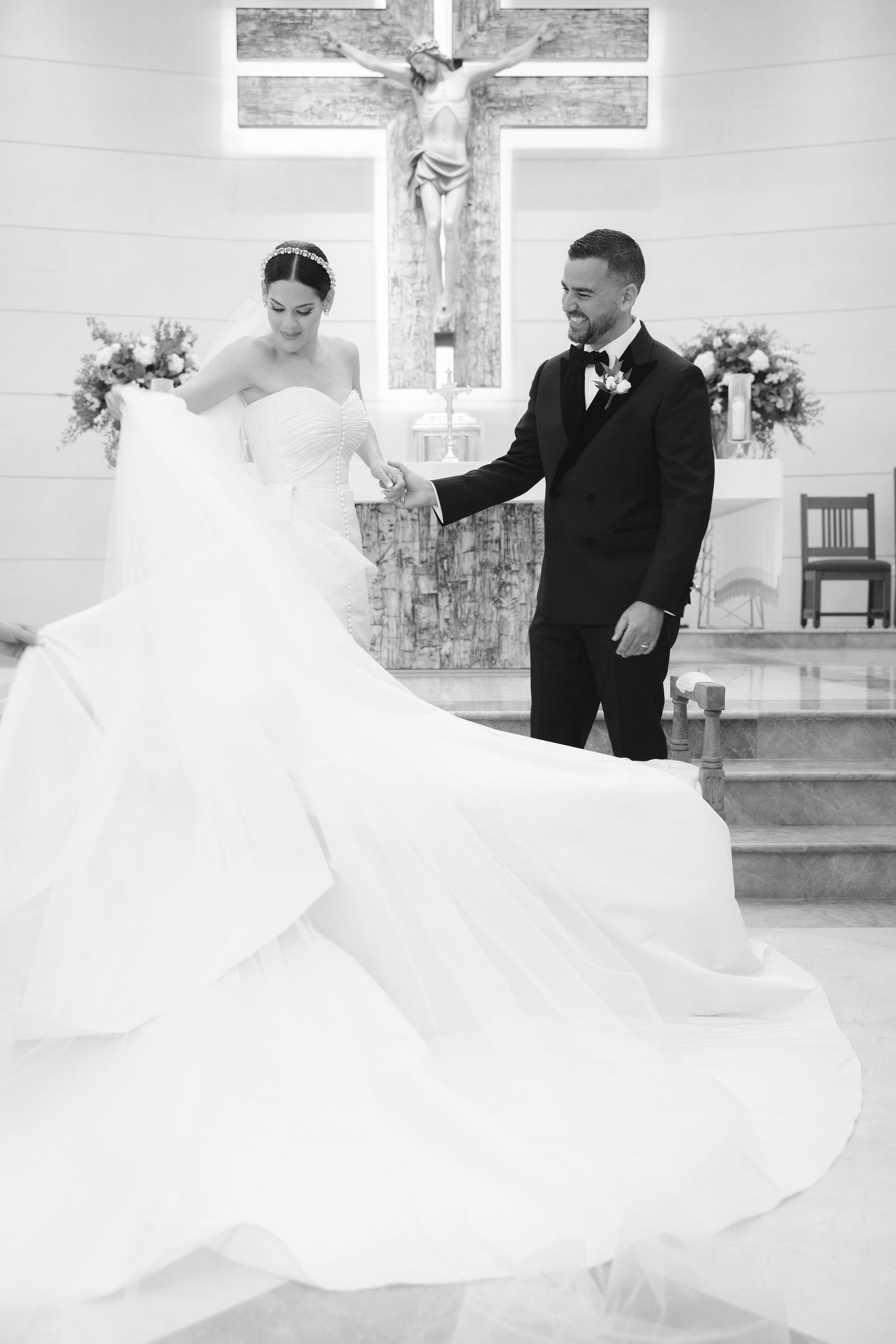 A black and white wedding photo showing a couple at the altar with a large crucifix and floral arrangements in a church.