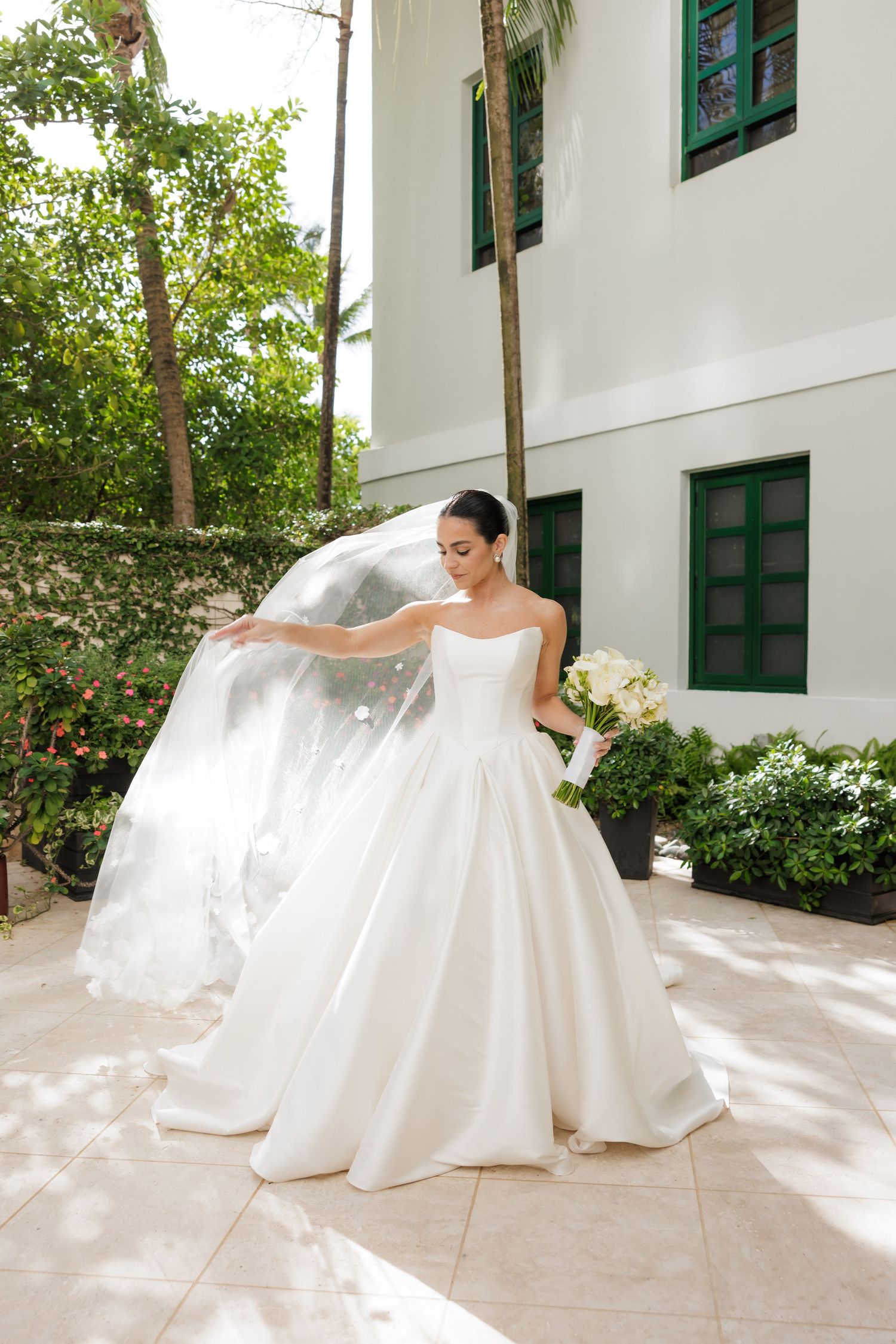 A bride in a stunning white ball gown poses with her flowing veil in a sunlit outdoor courtyard.