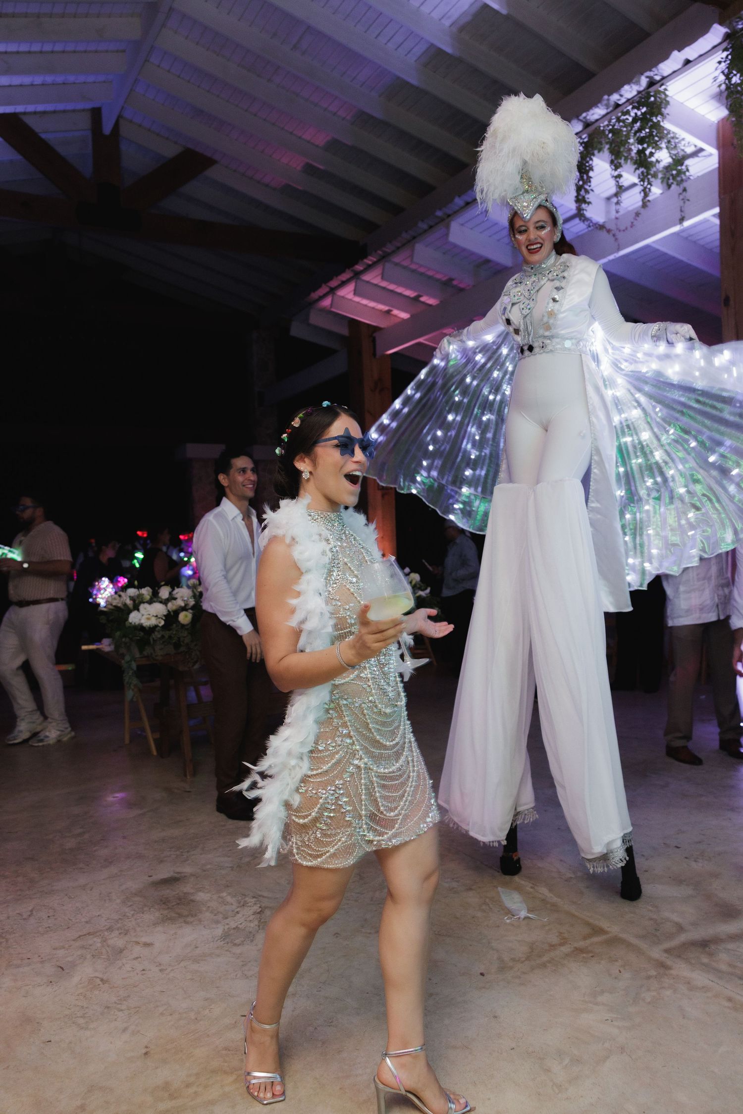Party guests dancing with illuminated LED wings under purple venue lighting.