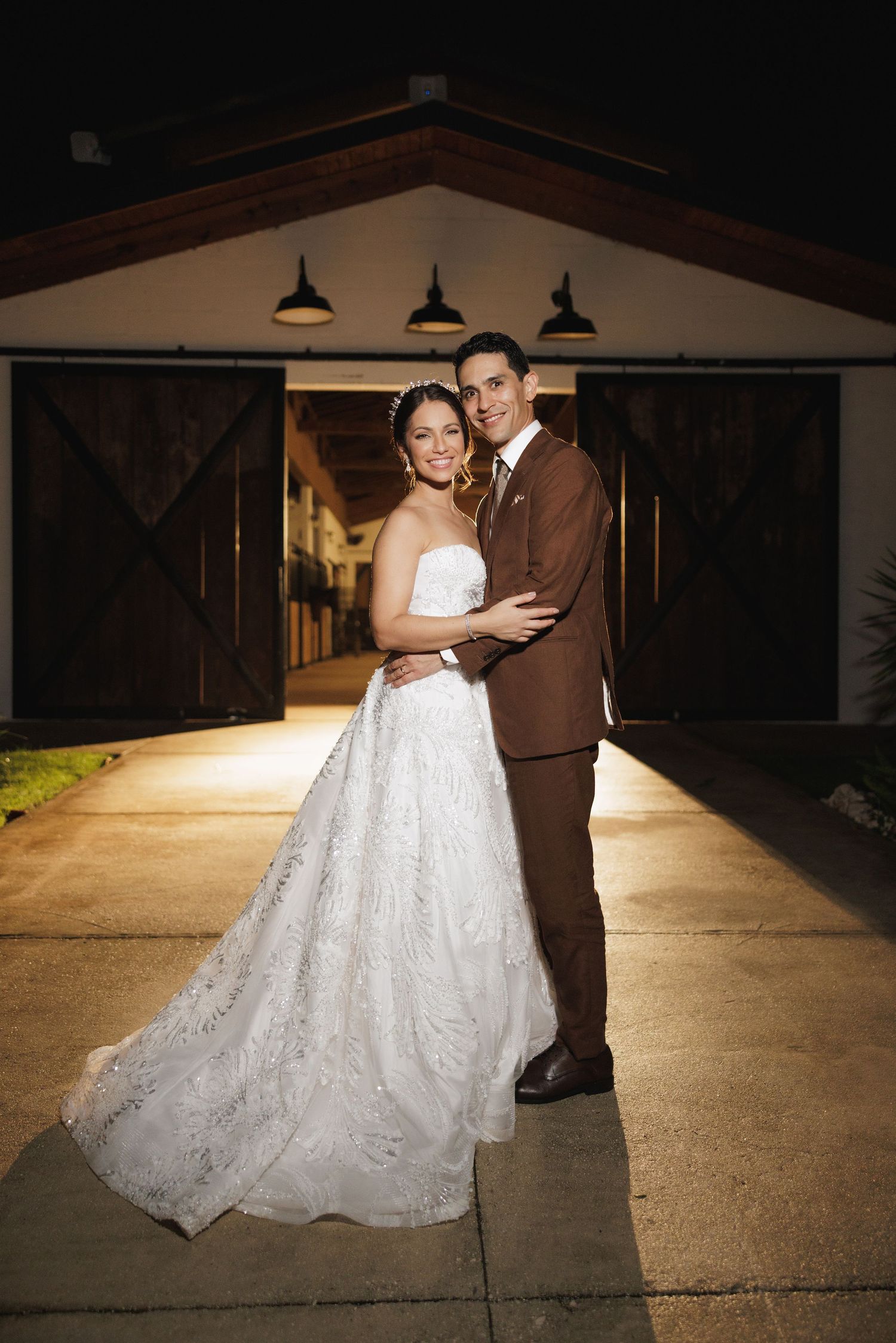 A newlywed couple embraces in front of a warmly lit barn entrance at night.