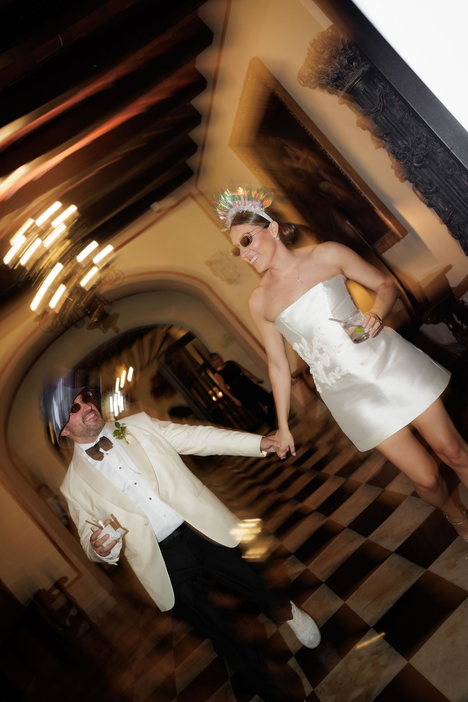 Dancing figures in white outfits perform energetic moves on a black and white checkered floor under chandelier lighting.