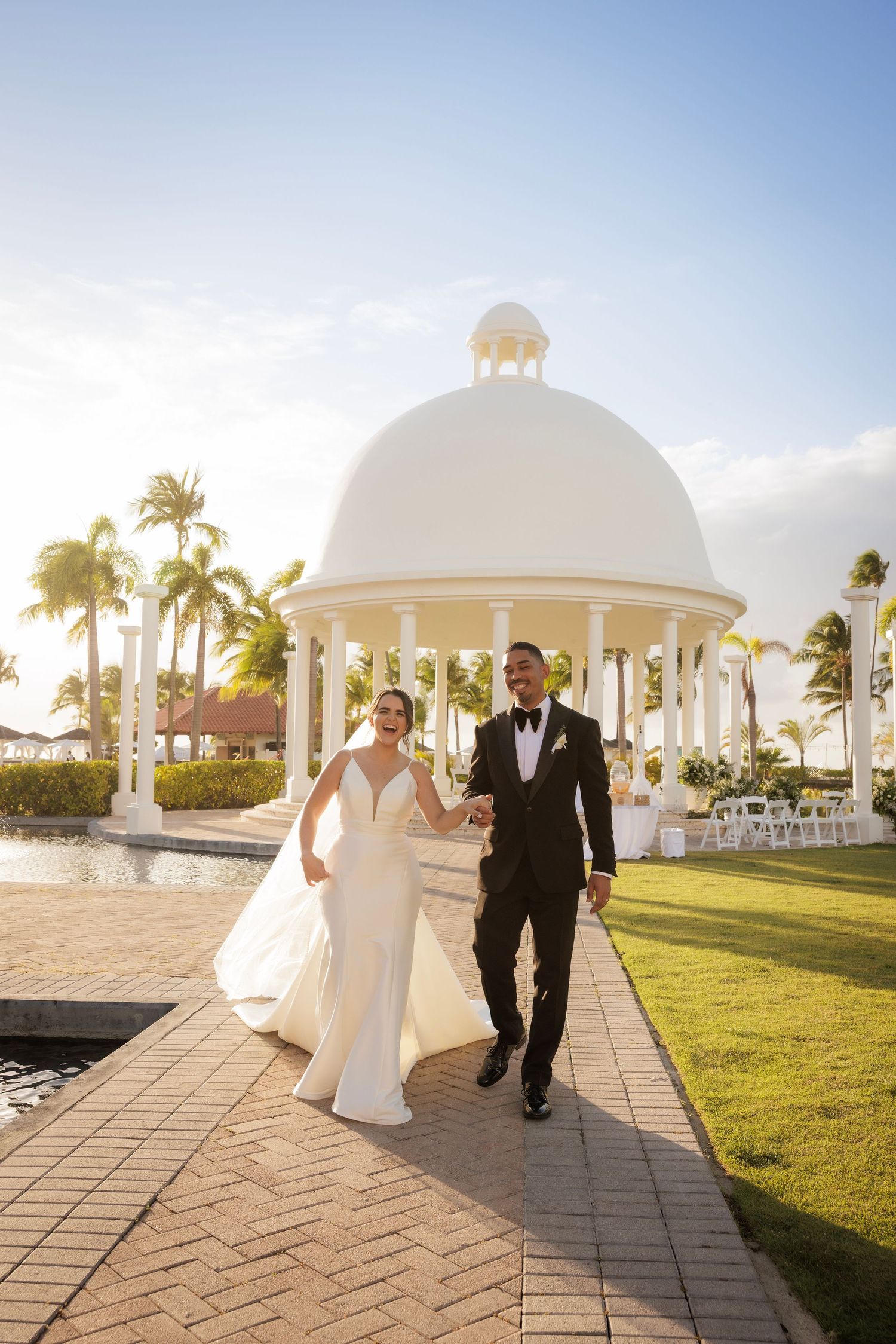A lovely panorama of a beachside wedding ceremony venue with elegant white gazebos and palm trees at sunset.