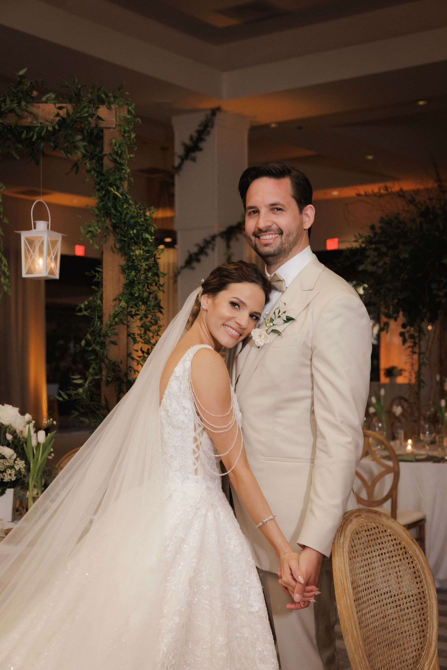 A bride in a lace wedding gown and groom in beige suit pose together in an elegant reception venue with greenery decor.