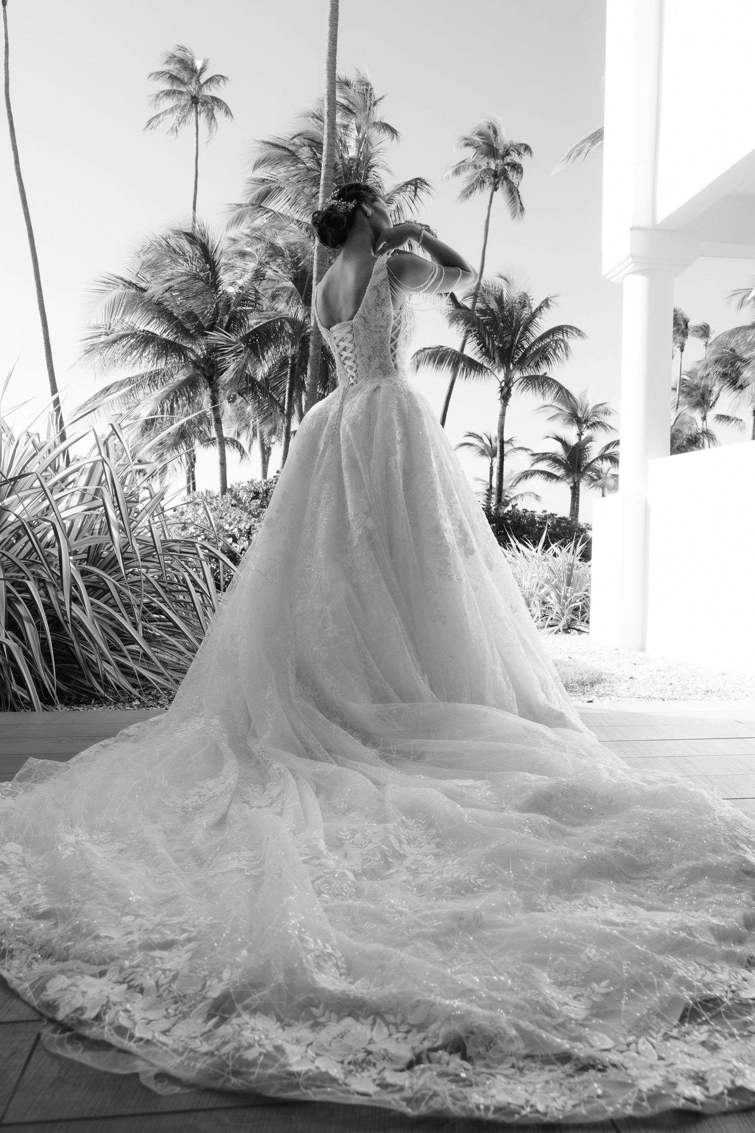 Black and white photo of a wedding dress with a flowing lace train against a backdrop of tropical palm trees and beach.