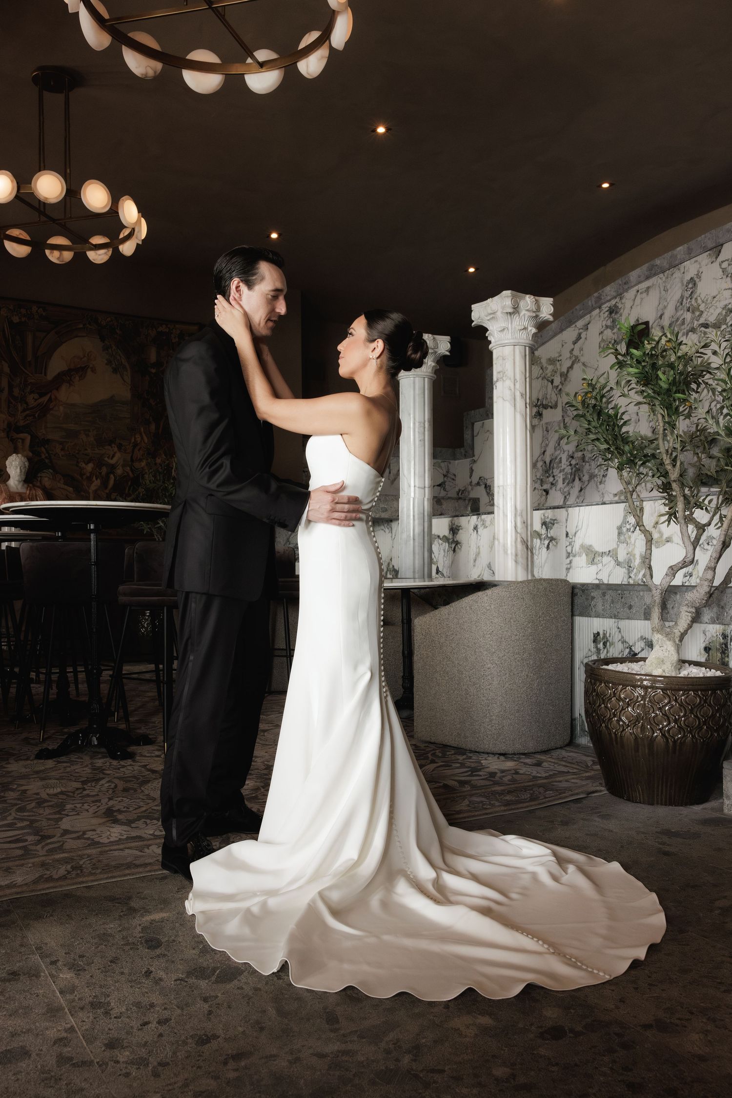 A newly married couple shares a romantic dance in a dimly lit reception venue with white columns and decorative lighting.
