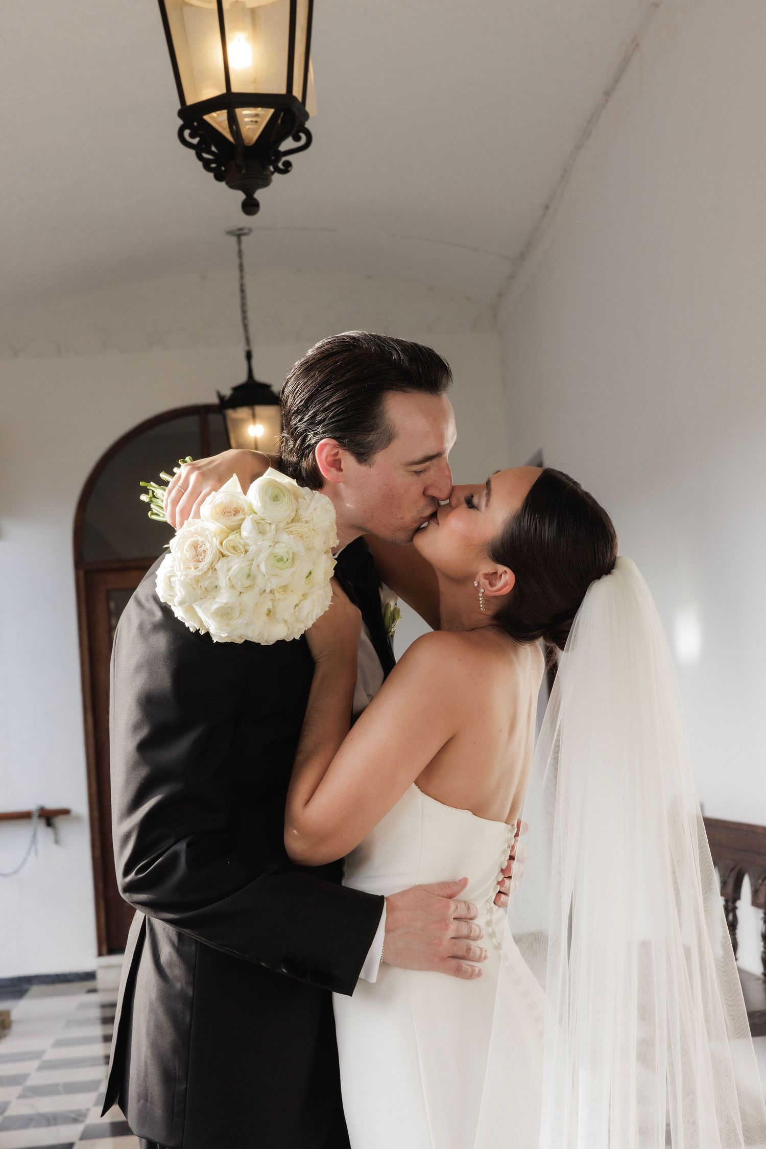 Romantic wedding kiss in an elegant white hallway with a black and white checkered floor.