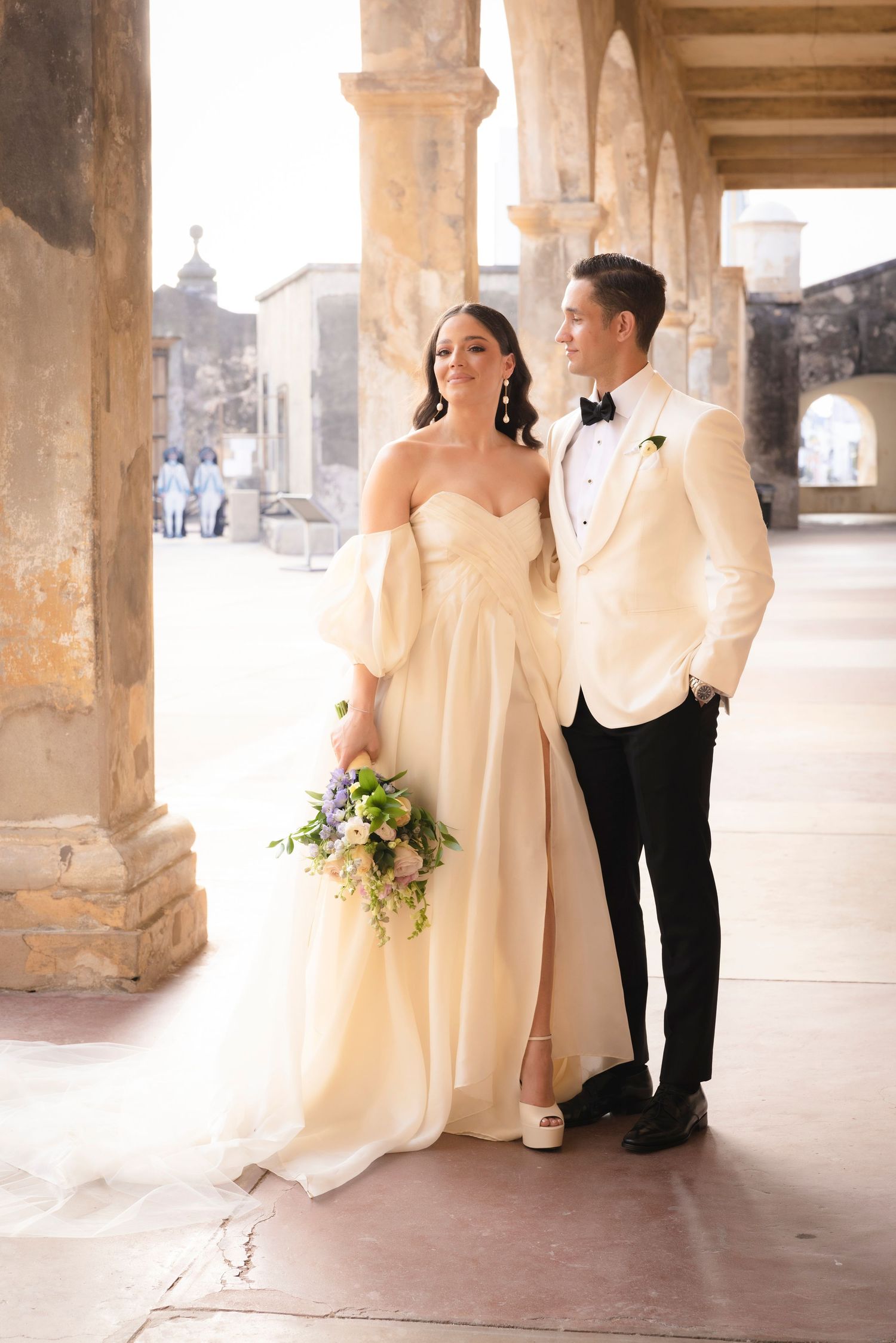 Couple in formal wedding attire poses under historic stone archways.