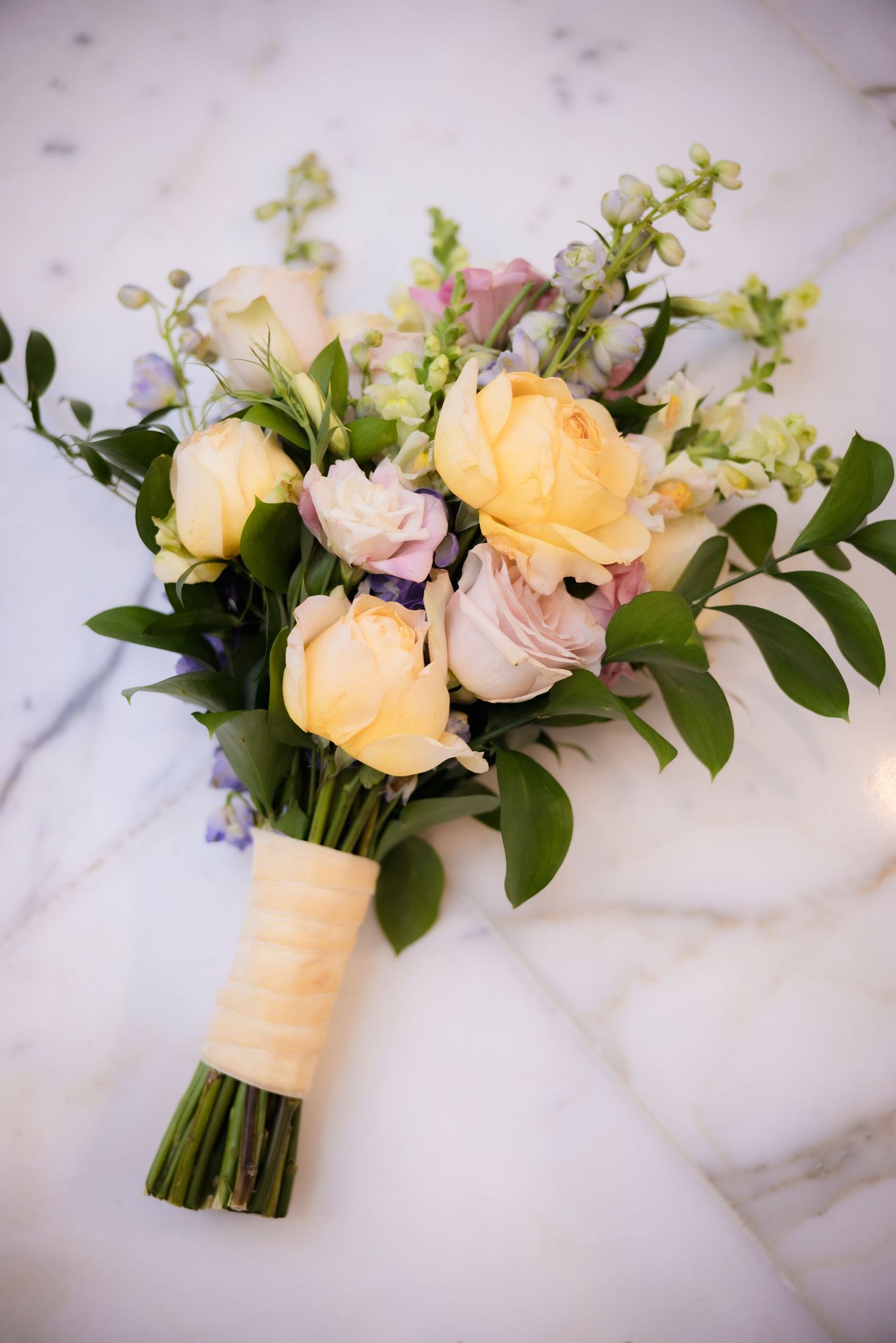 An elegant bridal bouquet featuring peach roses, pink flowers and greenery tied with a cream ribbon on marble surface.