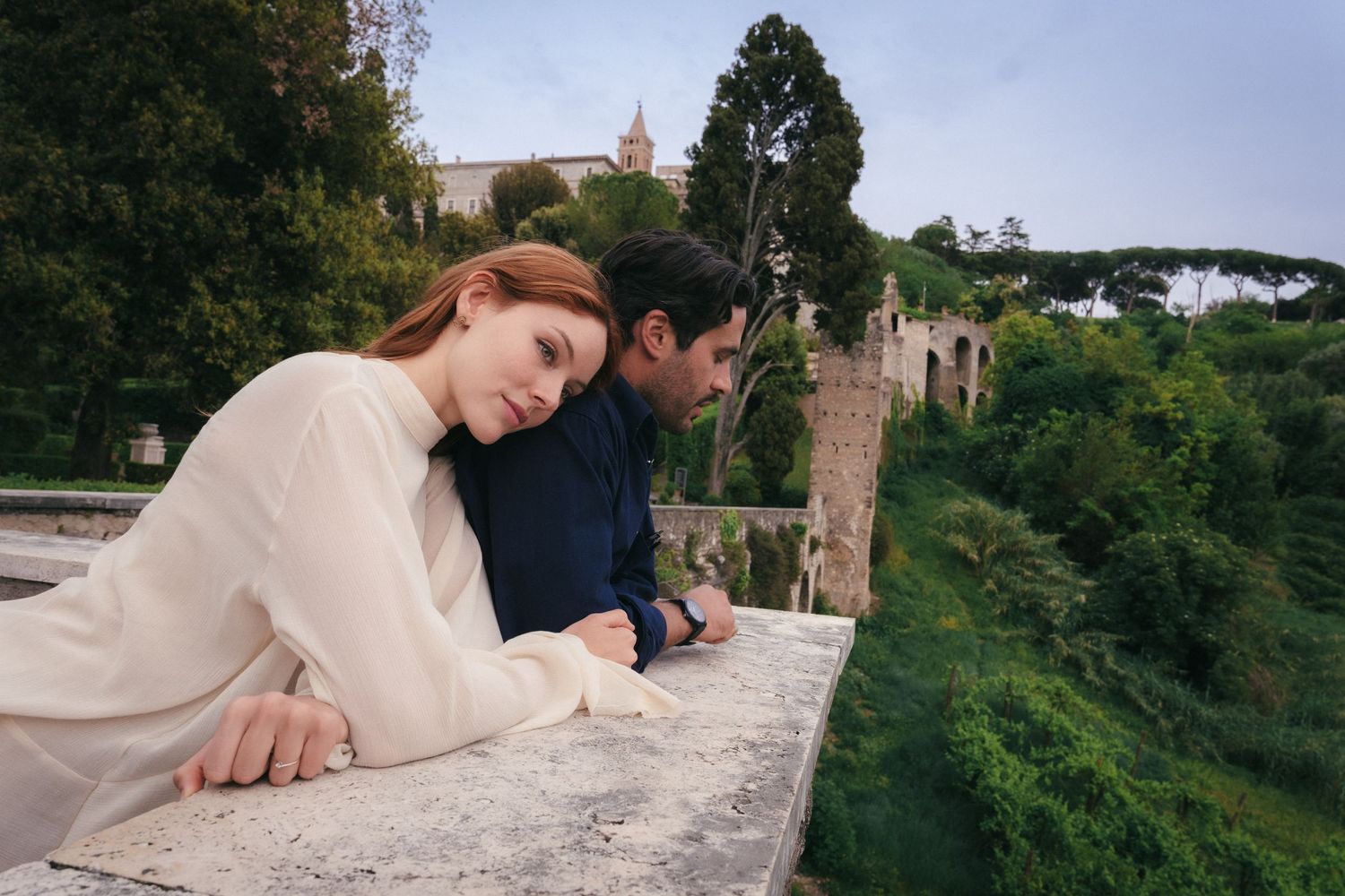 A couple leans on a stone balcony overlooking lush green gardens and ancient ruins in Rome, Italy.