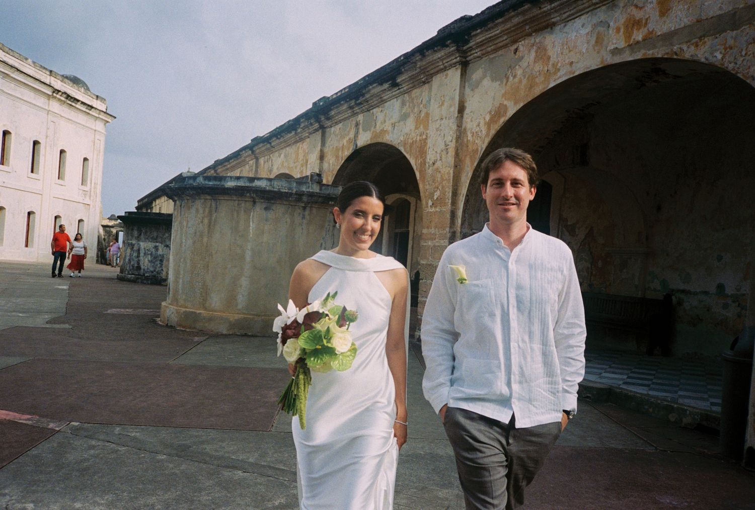 Bride in white gown holding bouquet walks with groom in white shirt near historic stone arches.