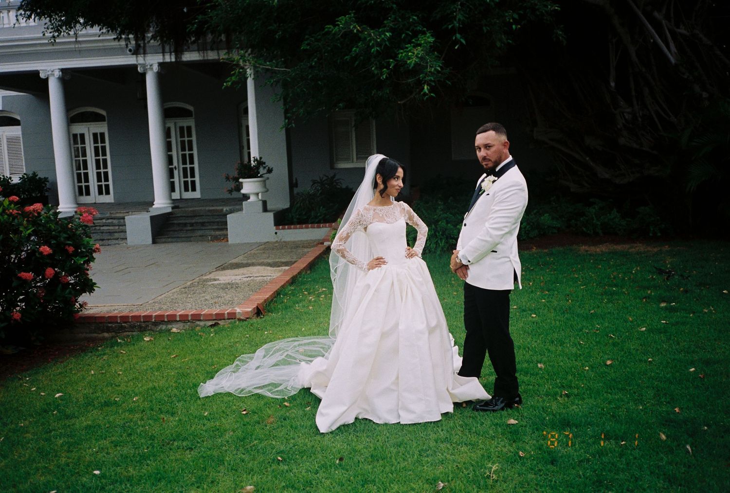 Bride in white ball gown and groom in white tuxedo posing on green lawn outside elegant Southern mansion.