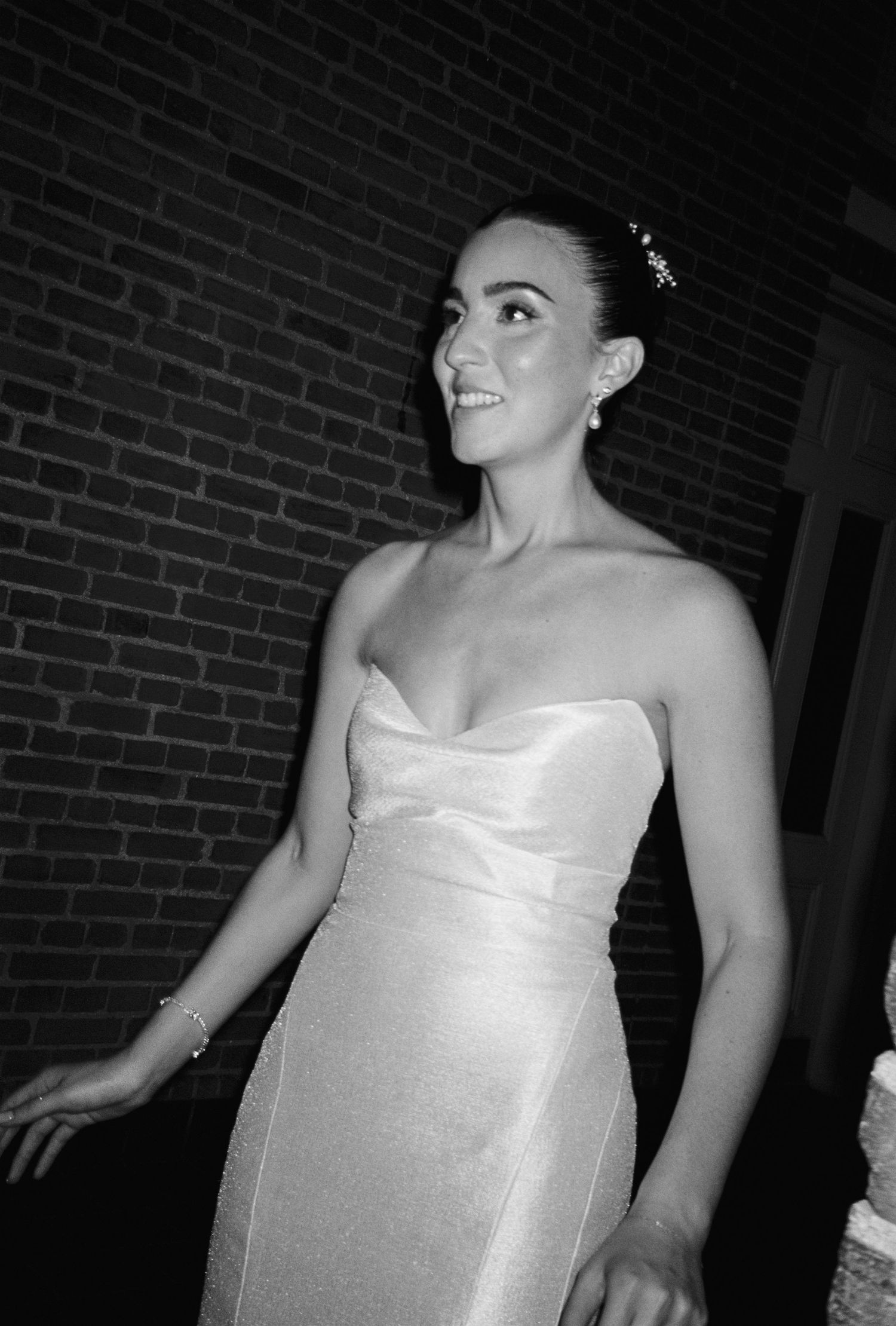 Bride in strapless white gown smiling joyfully in black and white photo against brick wall backdrop.