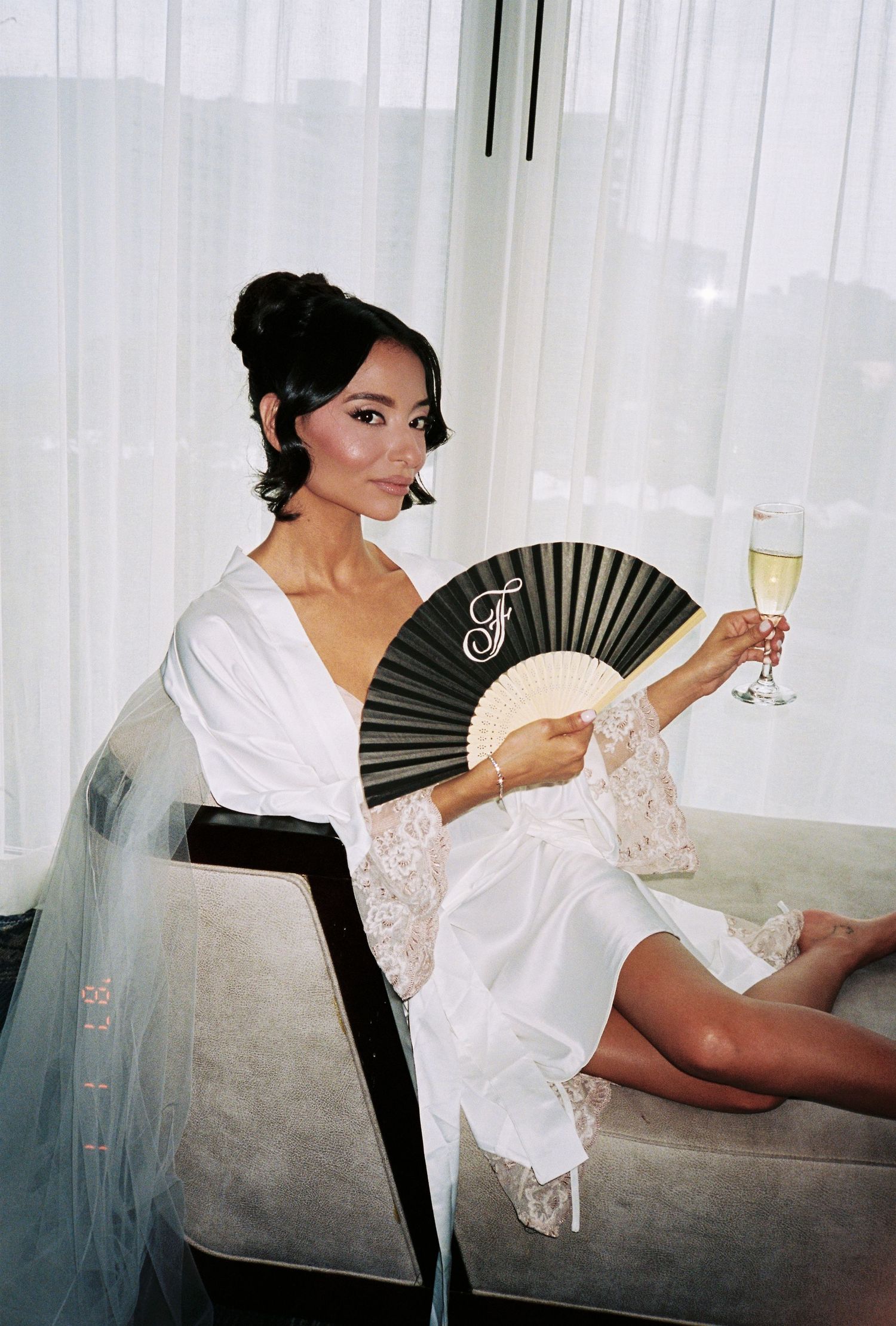 Bride in white lace robe holding a black monogrammed fan and champagne glass, seated elegantly before a window.