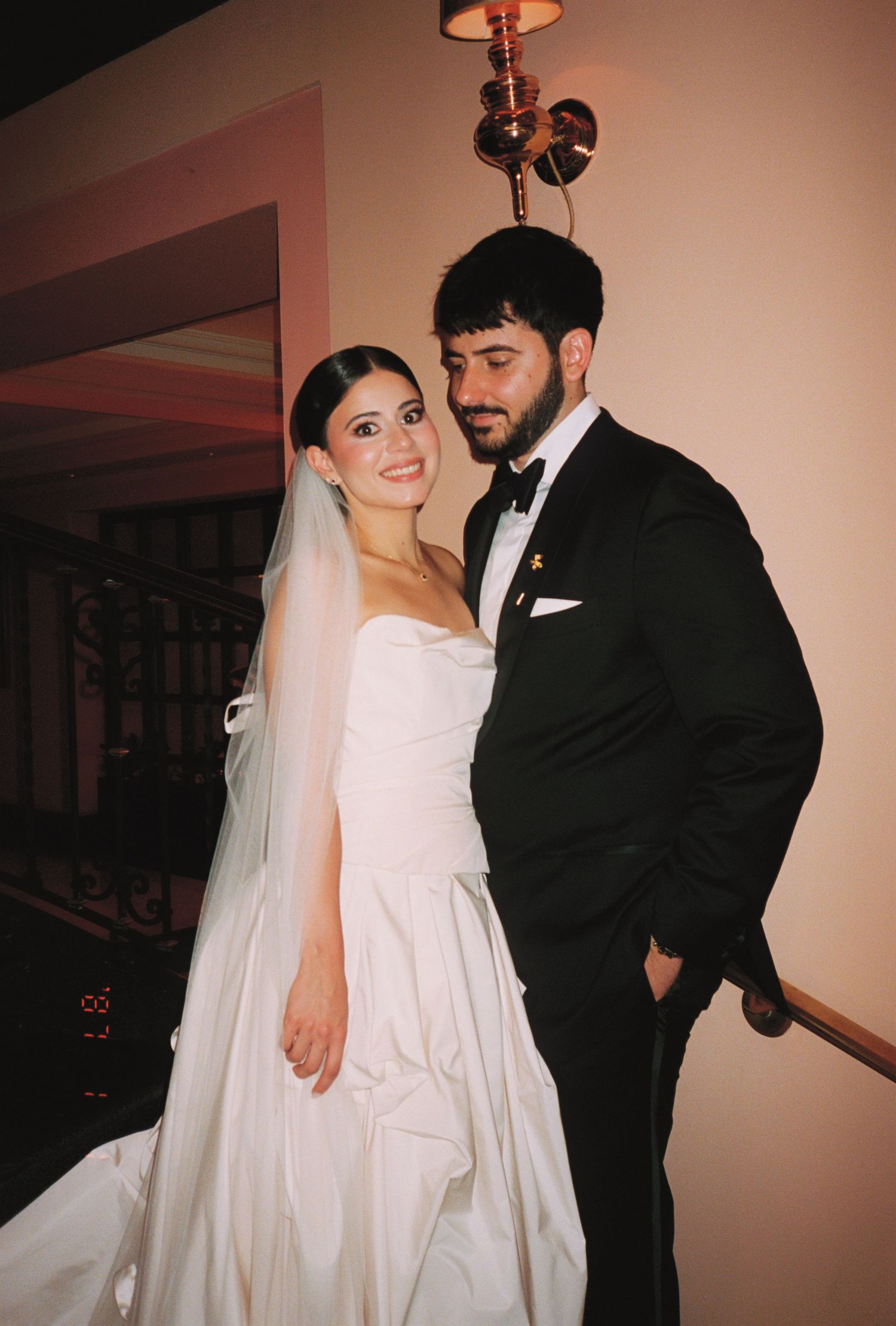 A bride in a white strapless gown and veil poses with a groom in a black tuxedo at their wedding reception.