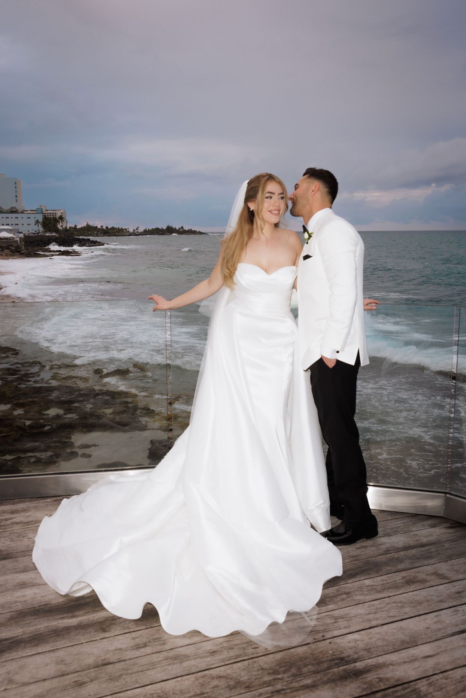 A romantic wedding couple poses on a wooden pier by the ocean as the bride's dress flows dramatically in the wind.