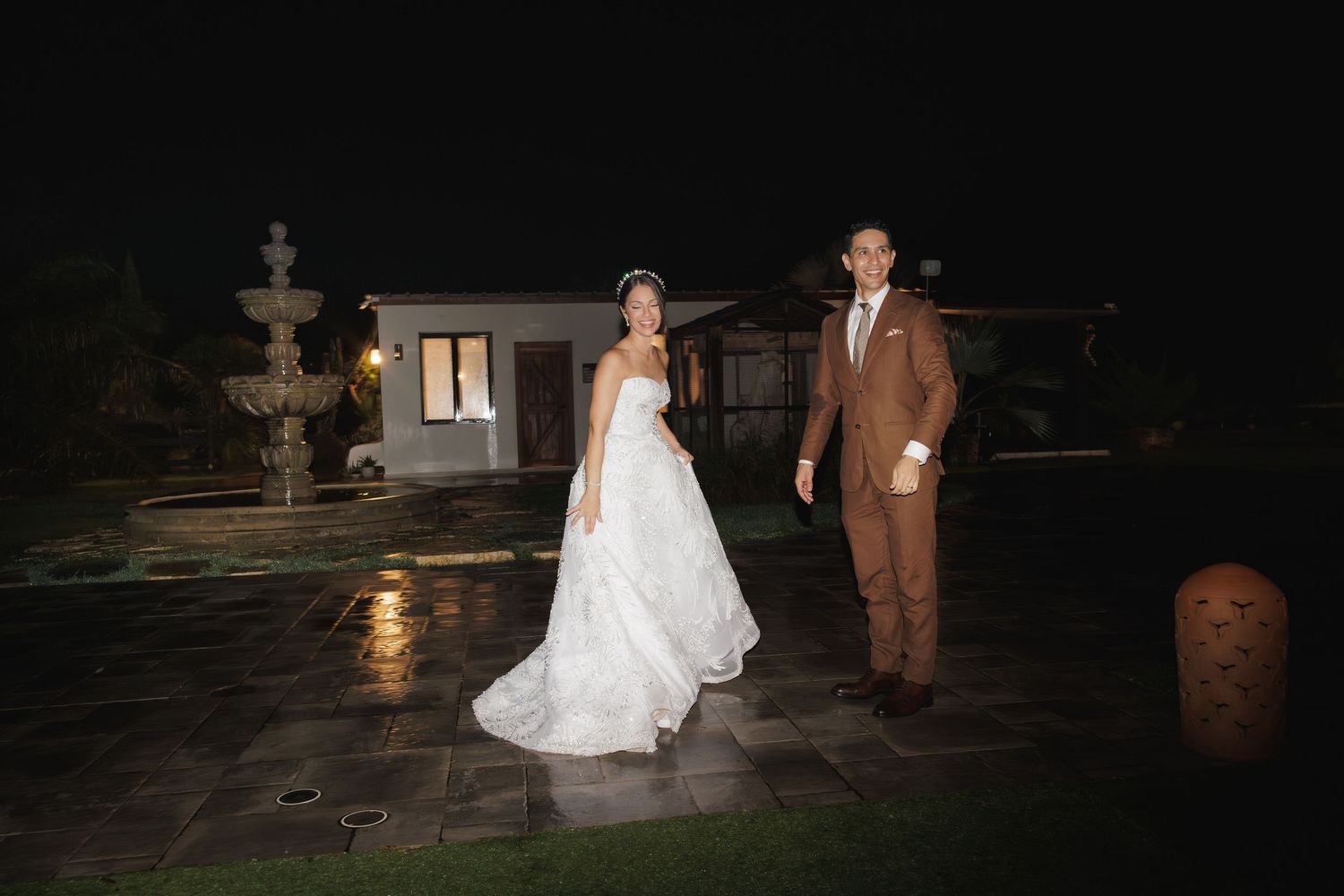 A bride in white gown and groom in brown suit share a romantic moment outside at night.
