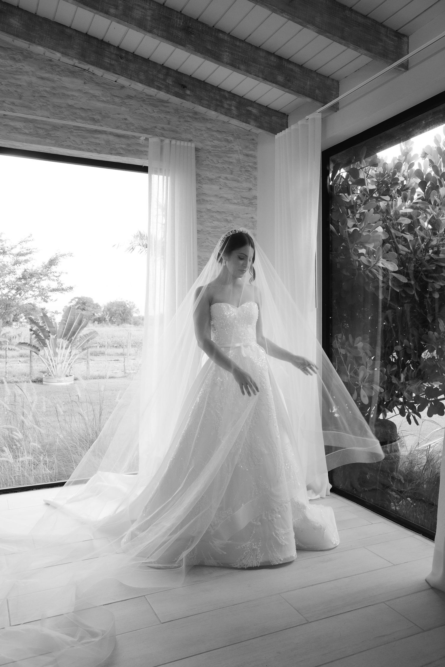 A black and white portrait of a bride in a flowing ballgown standing by large windows with natural light.