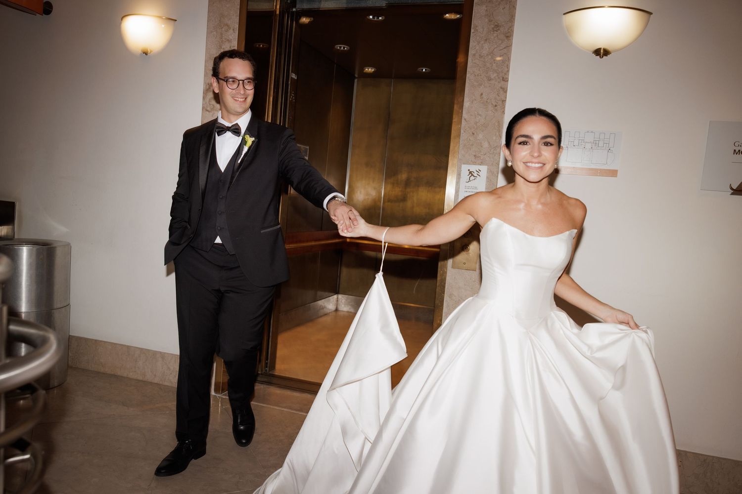A wedding couple and their guests walk down an elegant hallway with ornate lighting fixtures.