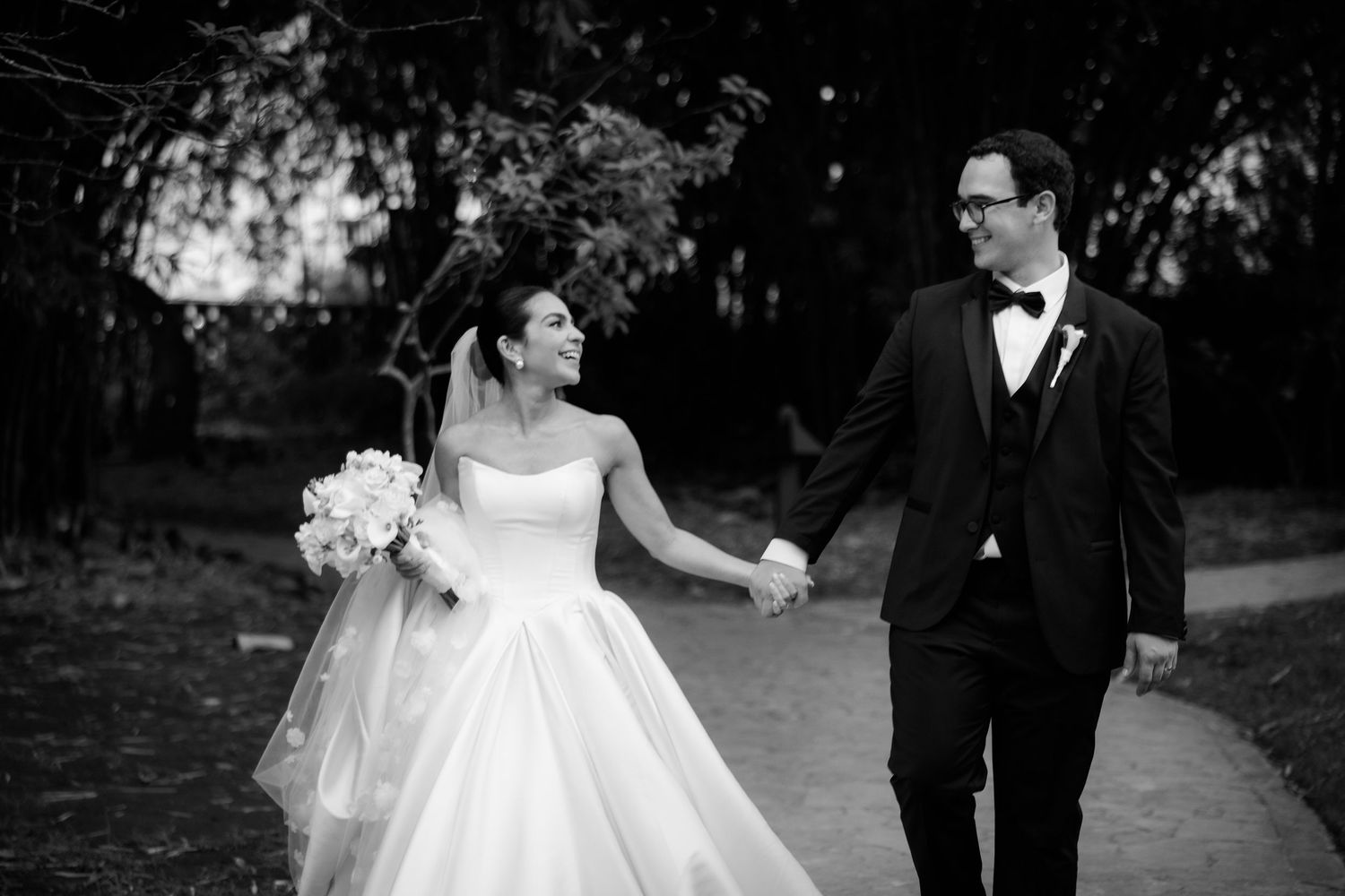 A bride and groom hold hands while walking together outdoors in a black and white wedding photograph.