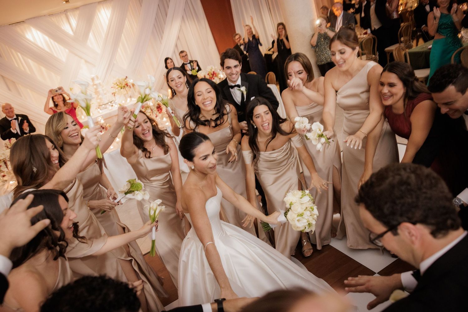Wedding party celebrates on the dance floor with bridesmaids in champagne dresses and white floral bouquets at reception.