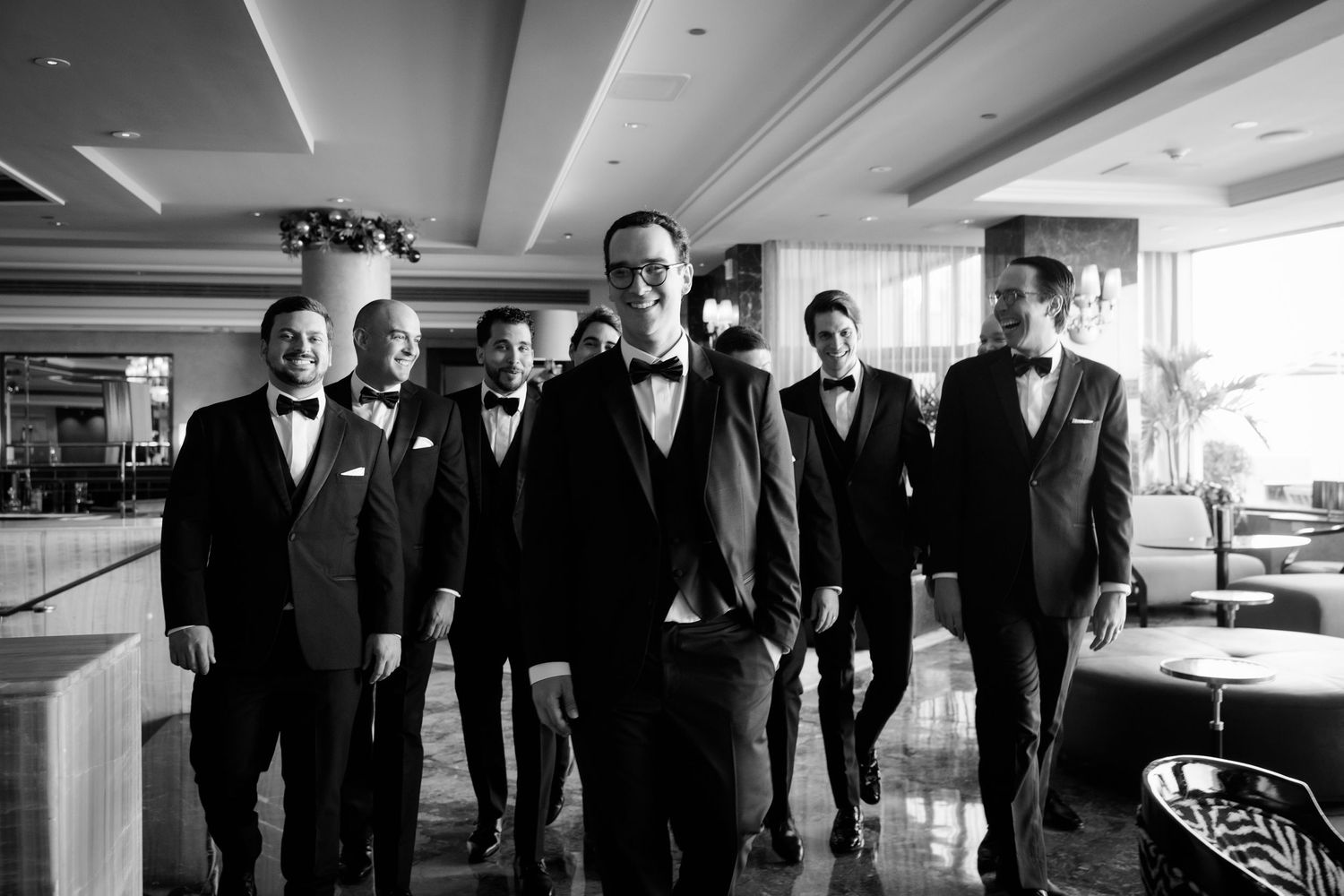 A black and white photo of groomsmen in tuxedos standing together in an elegant hotel lobby.