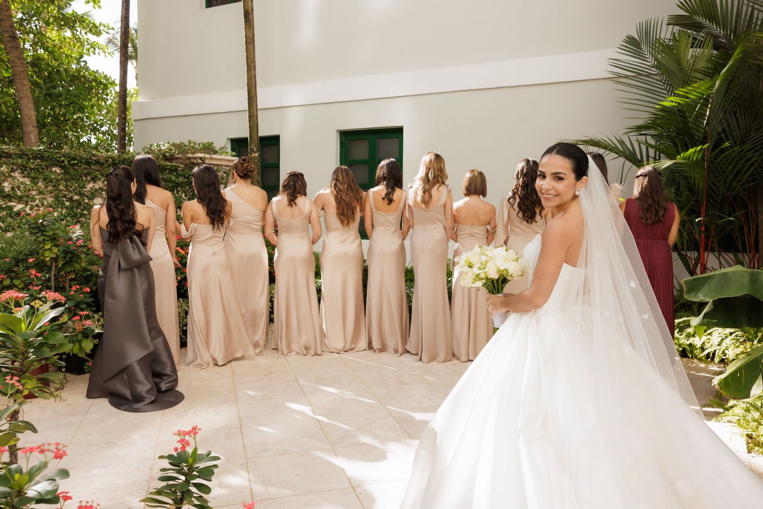 A wedding party poses together in a garden setting wearing blush pink bridesmaid dresses and a white bridal gown.