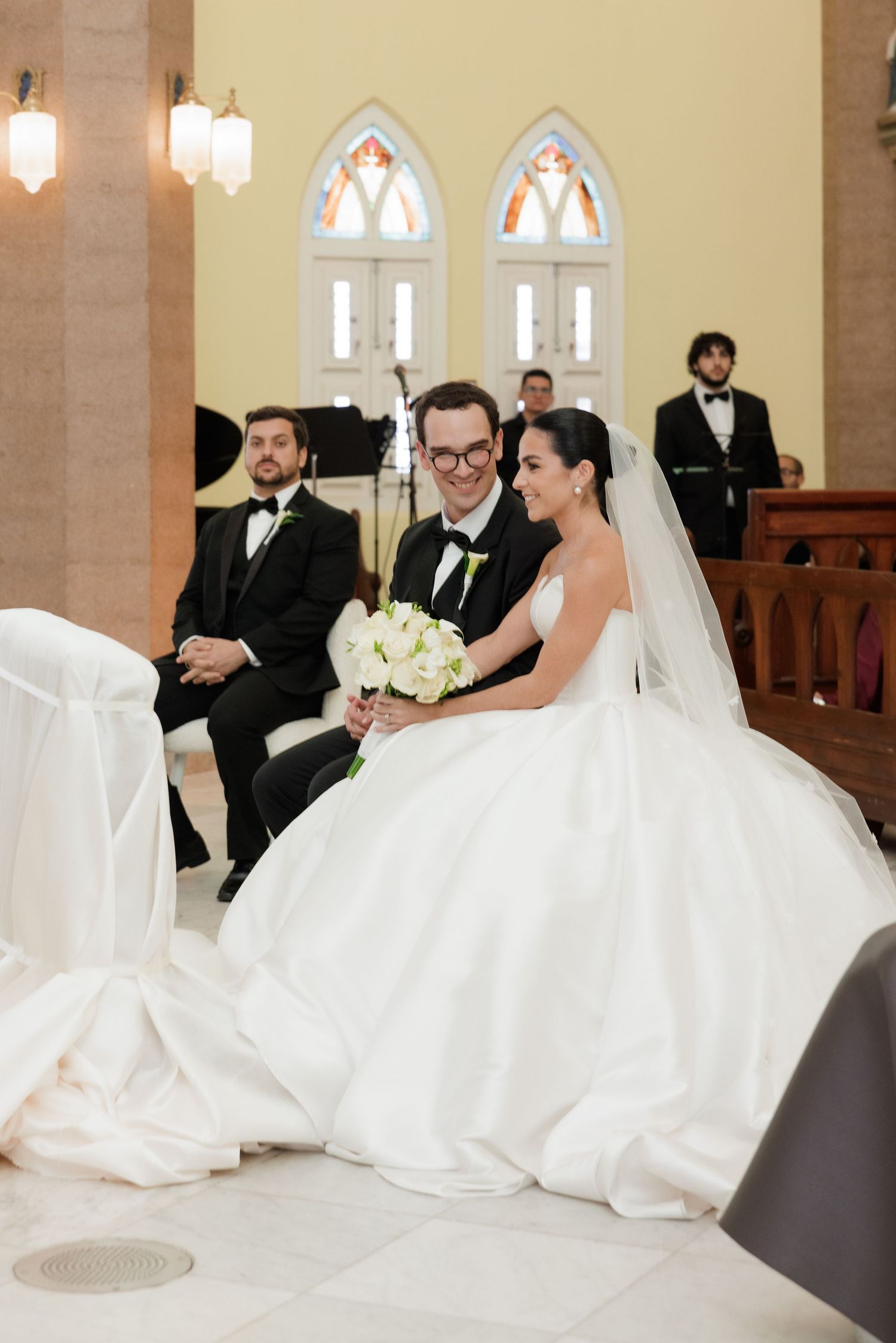 A wedding ceremony in a church with gothic arched windows and traditional wooden pews.