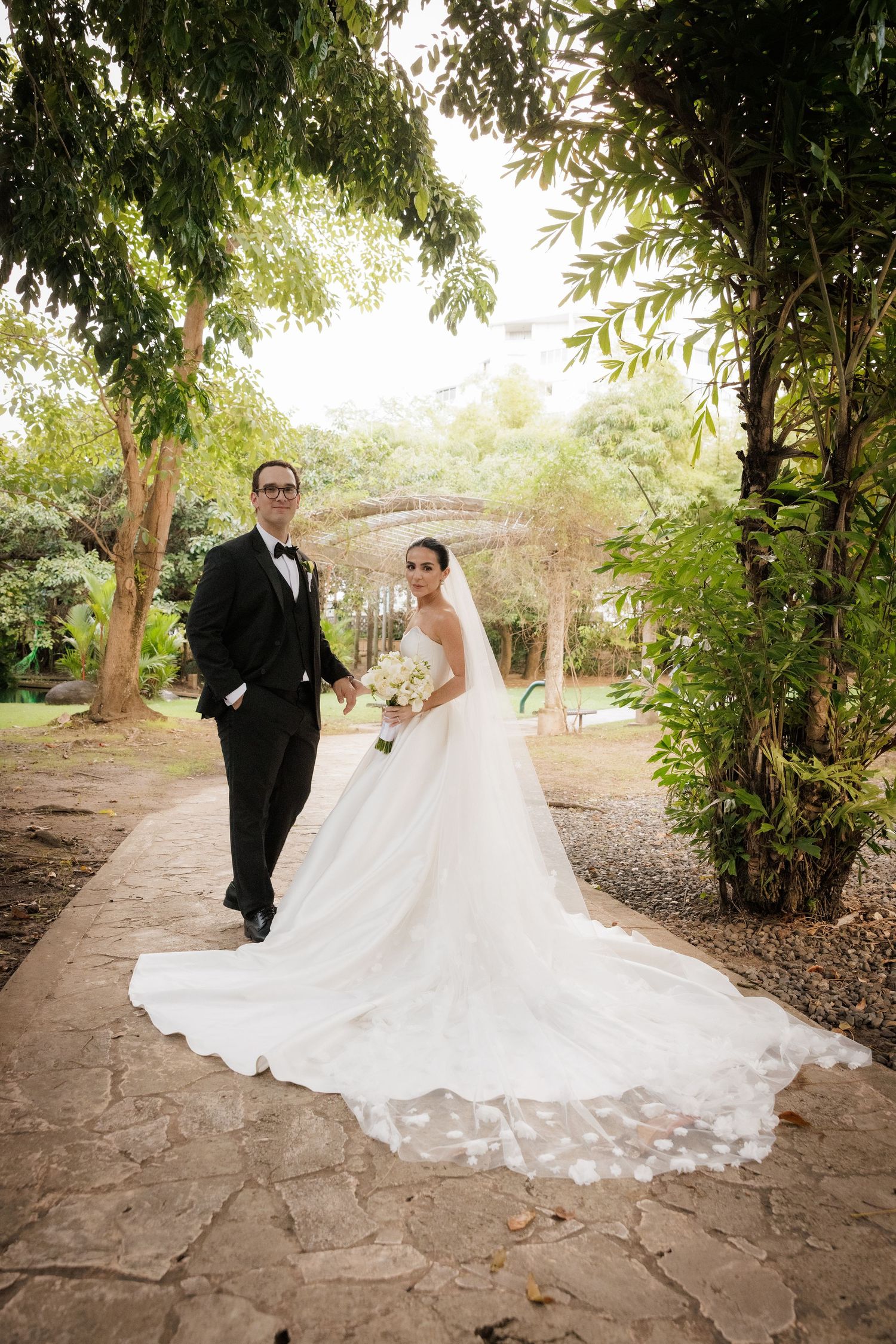 Wedding couple walking along stone path under lush green trees.