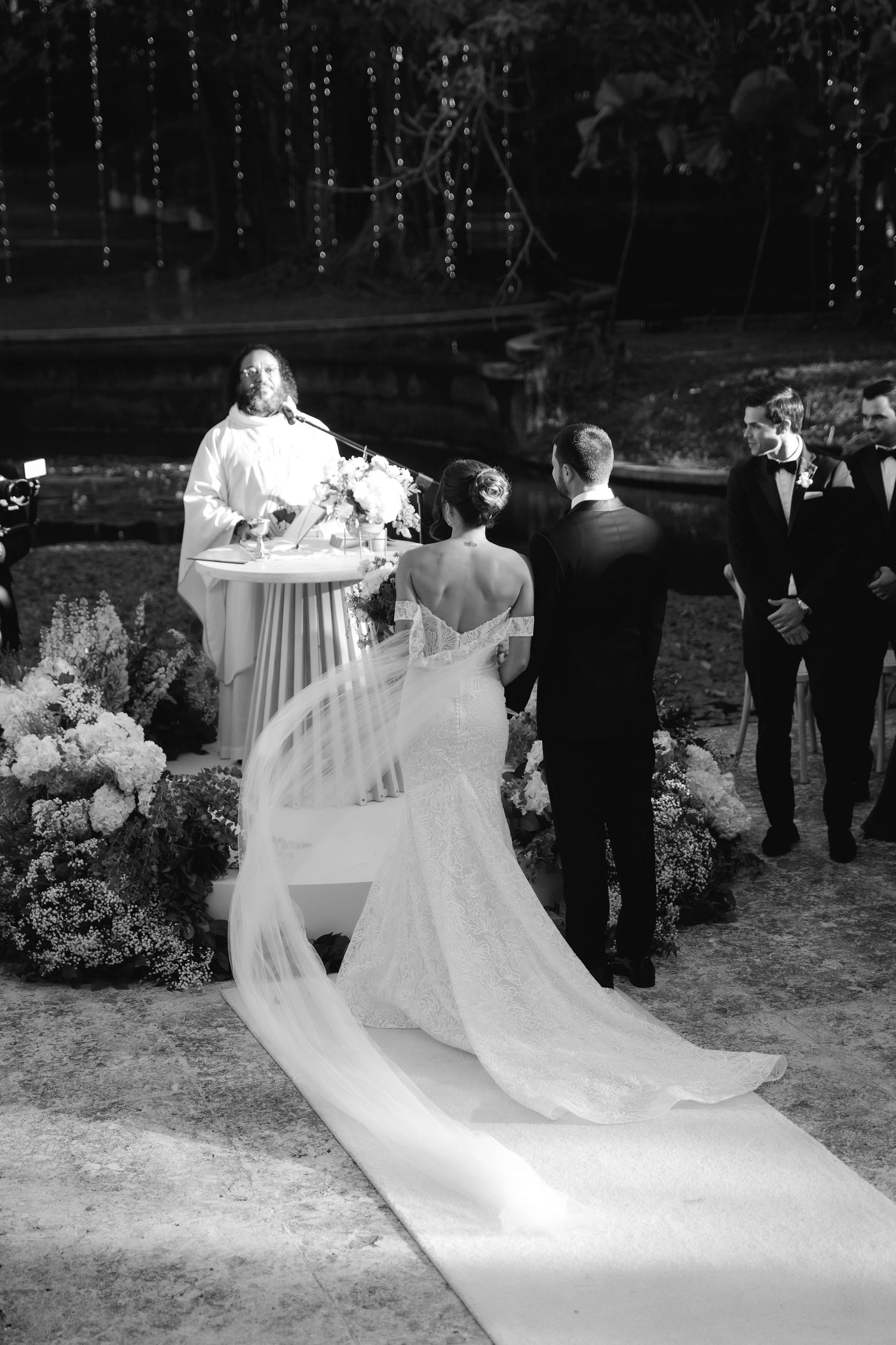 A black and white photo of a wedding ceremony with a long flowing veil and floral decorations under twinkling lights.