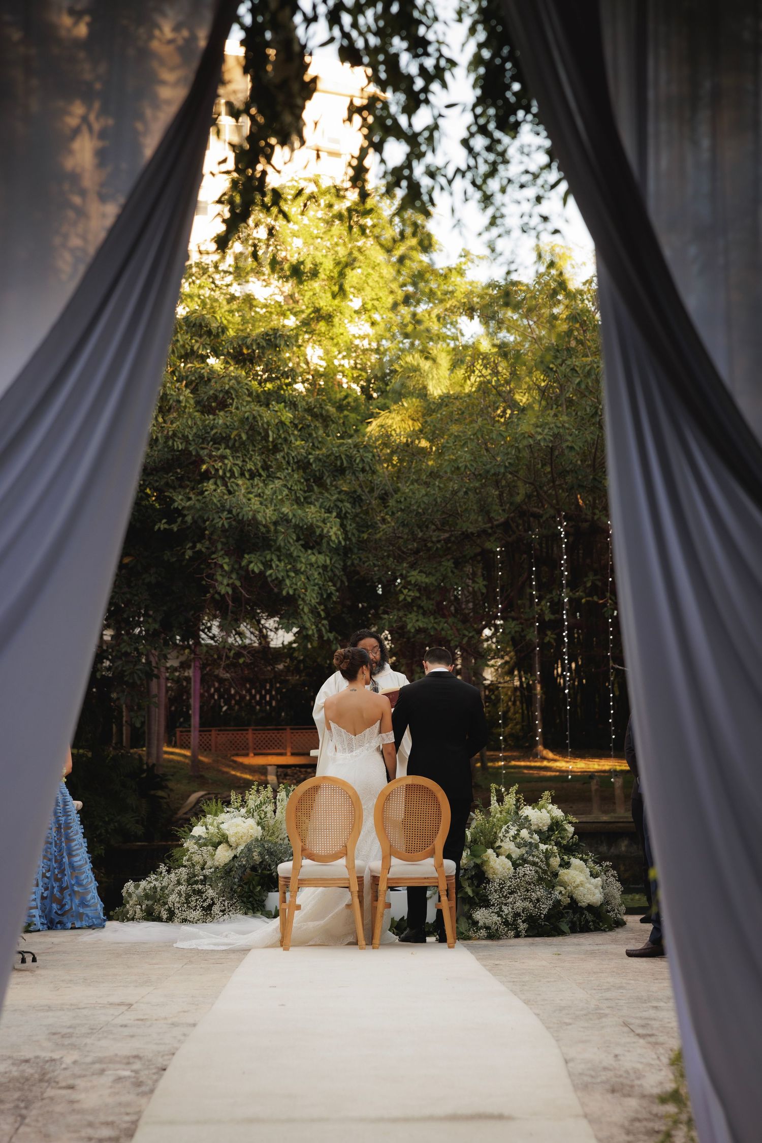 Couple sits at outdoor garden wedding venue framed by draped fabric and rattan chairs with white floral arrangements.