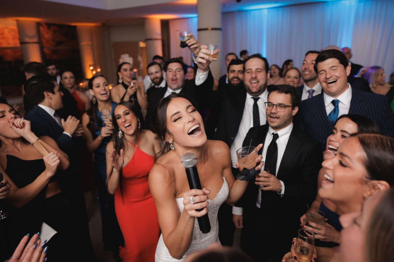 Wedding reception guests singing and laughing together on dance floor with drinks and festive party atmosphere.