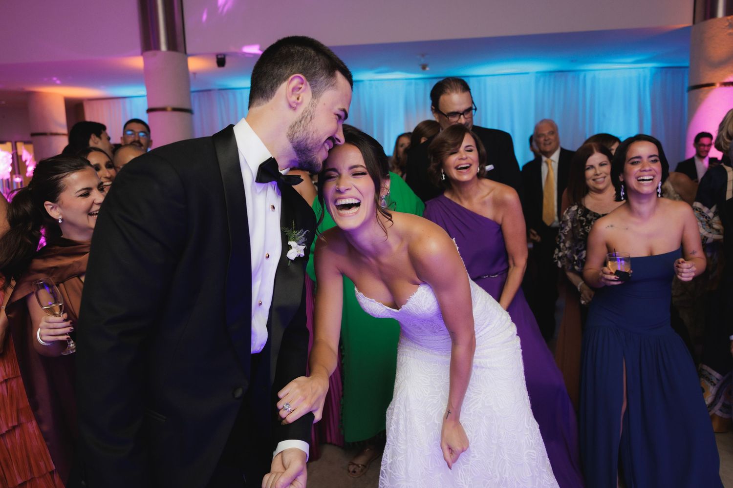 Wedding guests share a joyful moment on the dance floor at a reception with purple and blue lighting.