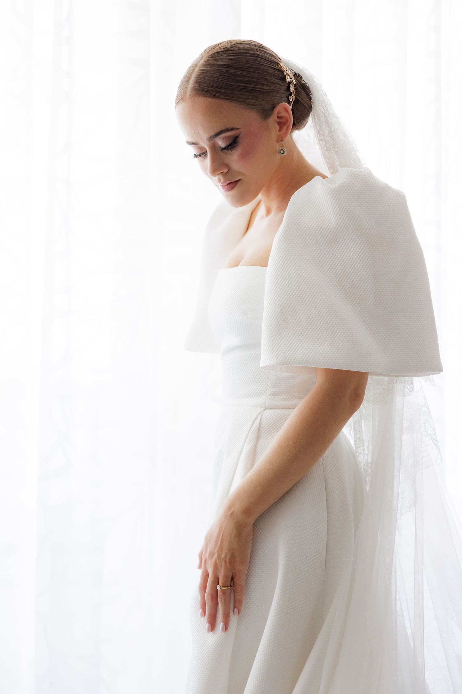 Elegant bride in modern minimalist wedding dress poses gracefully against white backdrop in artistic portrait series.
