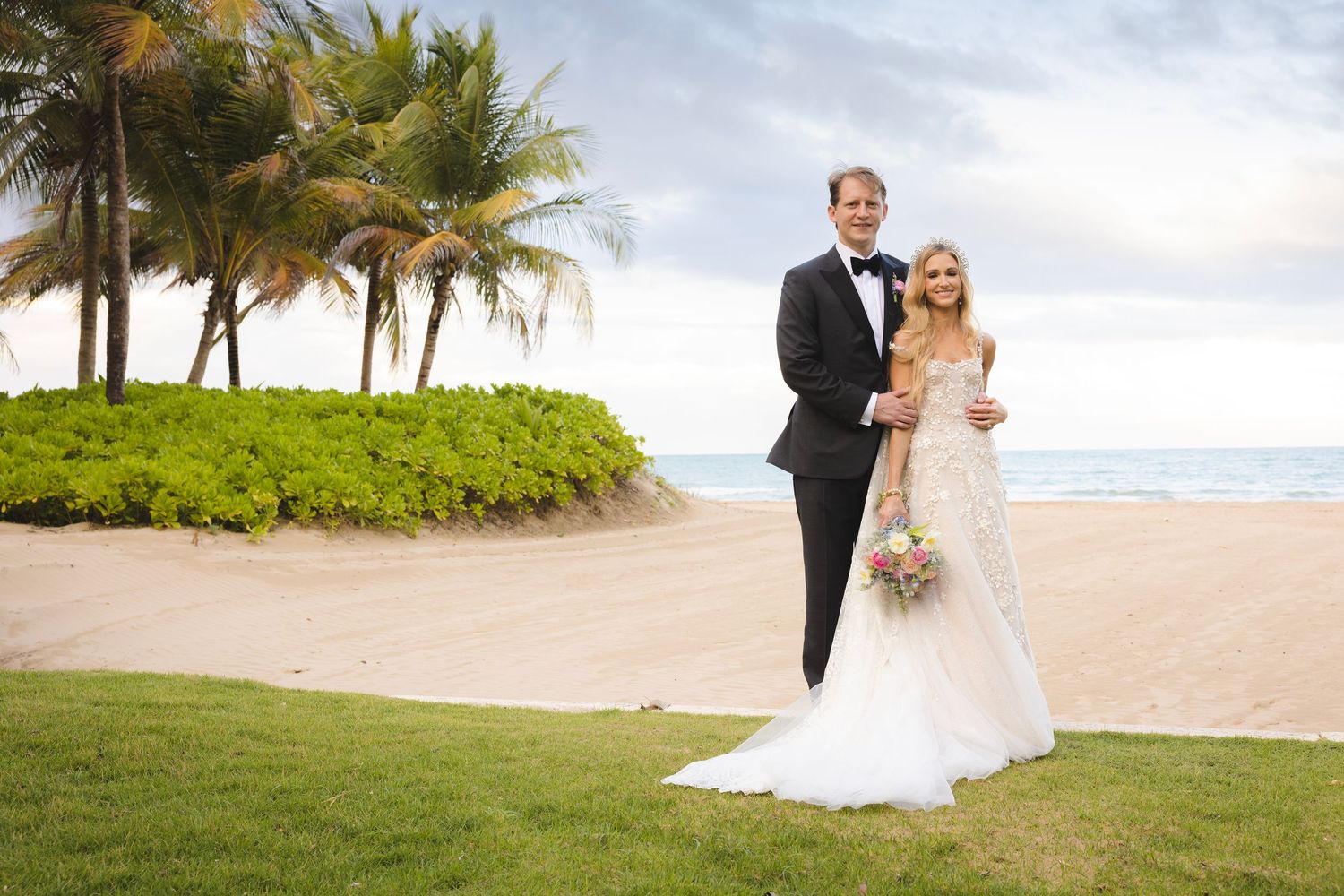 A couple in formal wedding attire stands on a scenic beach with palm trees and ocean views in the background.