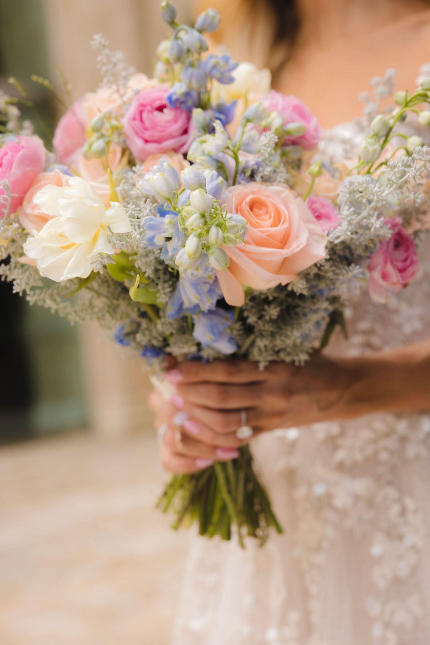 A romantic bouquet with pink and peach roses, blue delphiniums, and delicate baby's breath against a lace dress background.