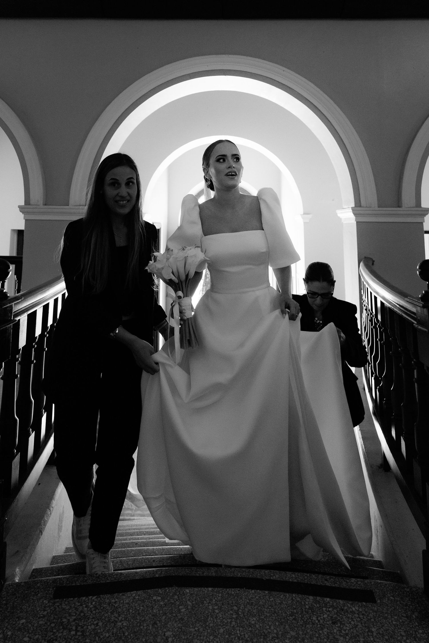 Black and white photo of a bride in a structured gown descending an arched staircase with assistance.
