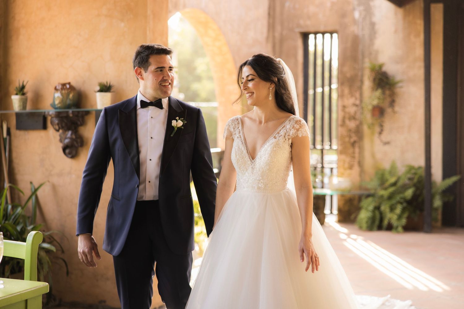 Elegantly dressed bride in white gown and groom in tuxedo walk together through warmly lit Mediterranean-style corridor.