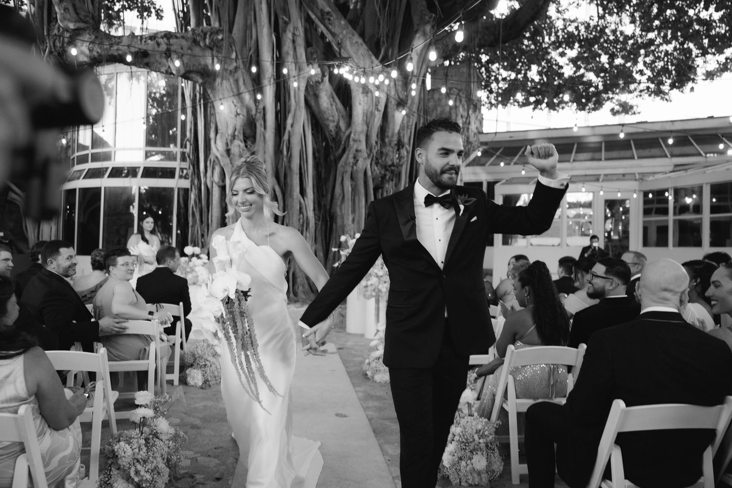 A joyful wedding couple exits their outdoor ceremony holding hands while guests watch from white chairs under a banyan tree.