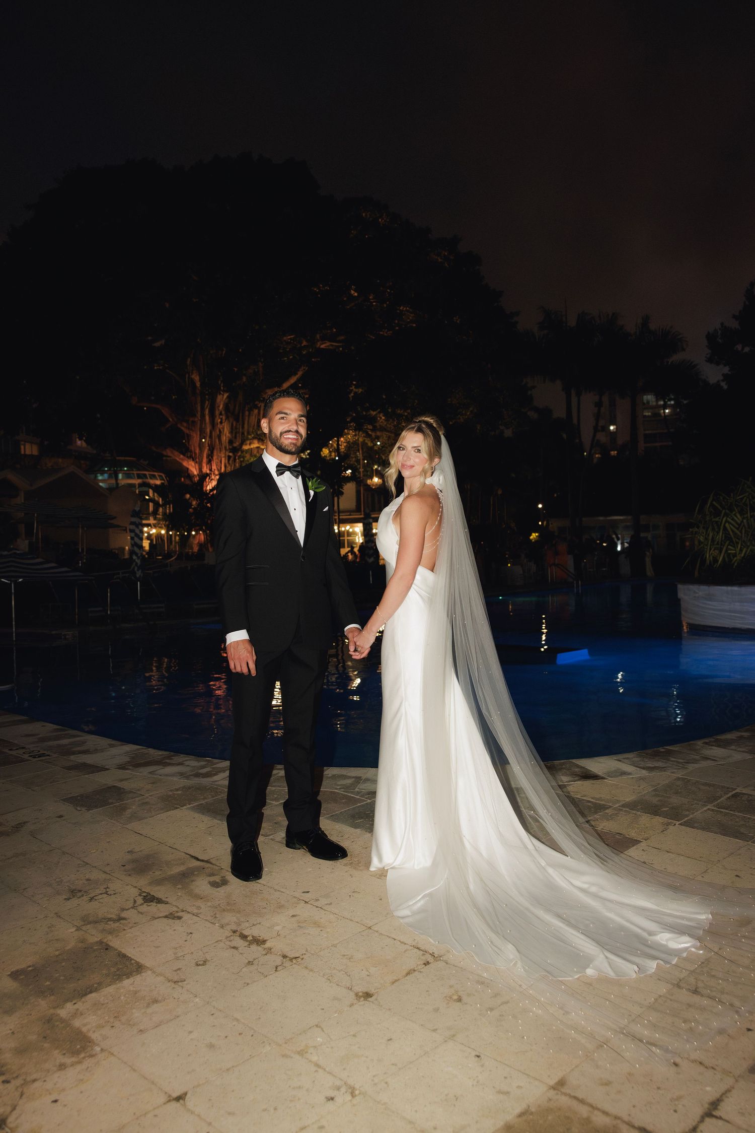 A couple stands together at night by an illuminated pool, with the bride wearing a flowing white gown and long veil.