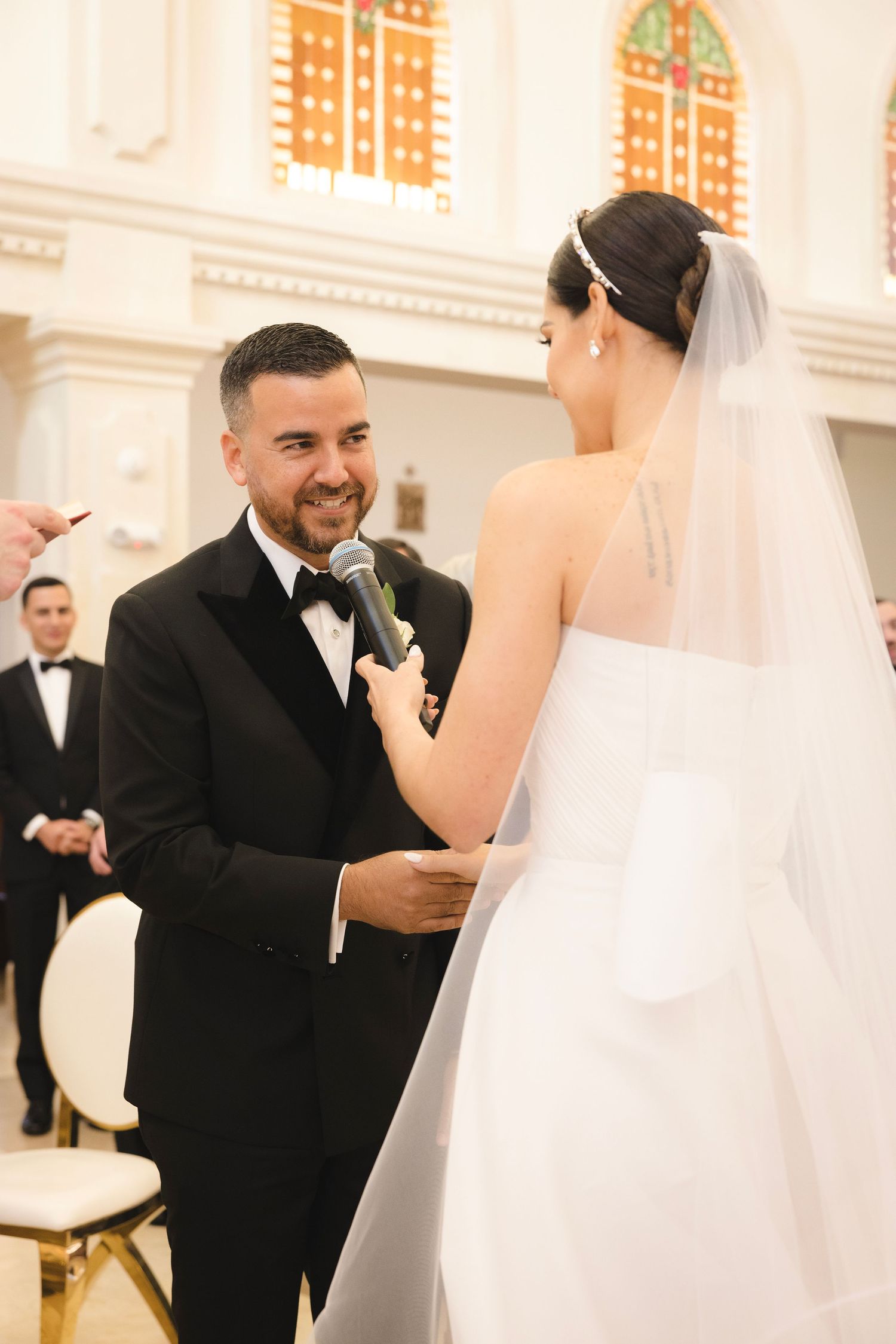 A couple exchanges loving glances during their wedding ceremony in an elegant white-walled room with stained glass windows.