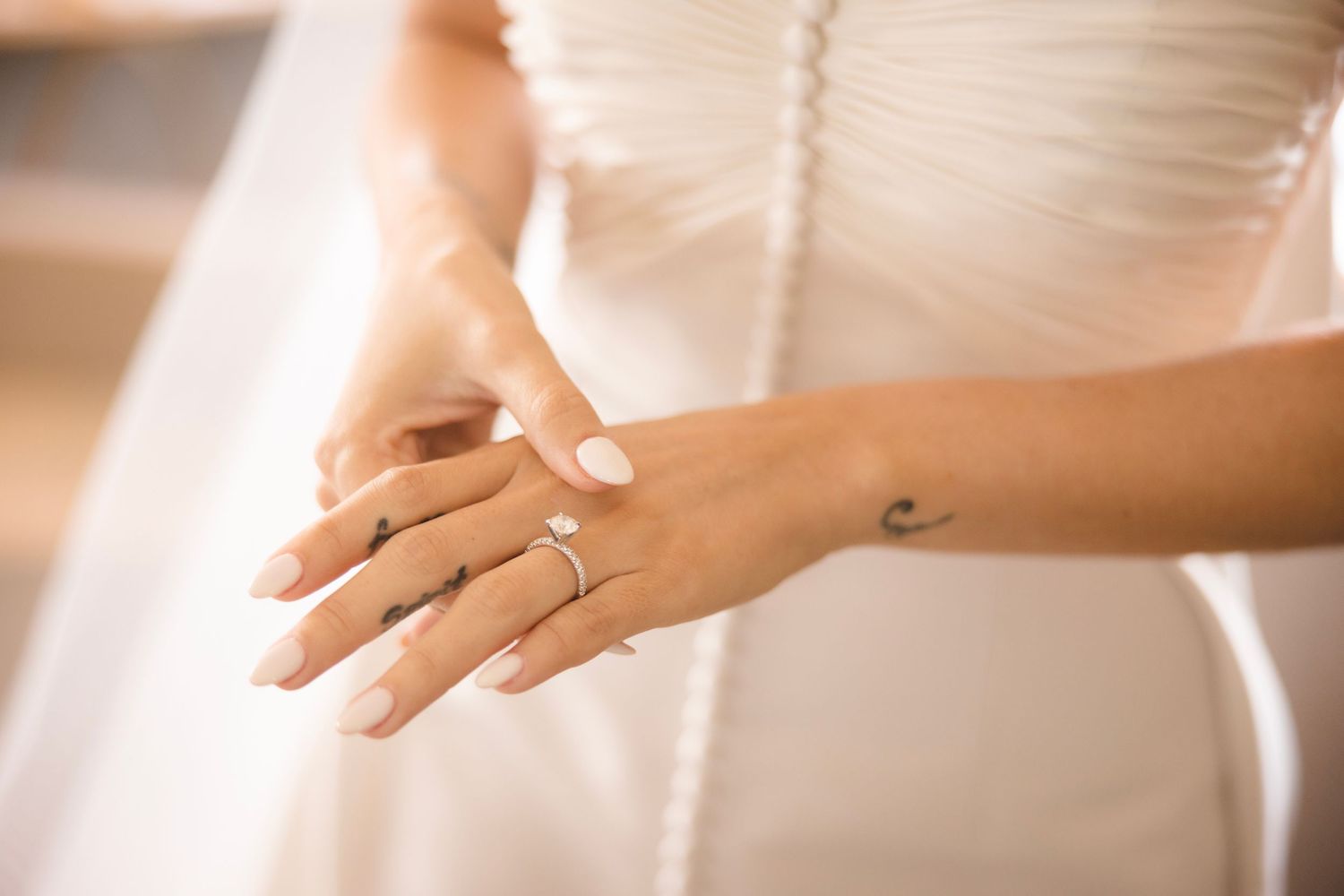 Close-up of bride's hand with engagement ring and white wedding dress in warm lighting.