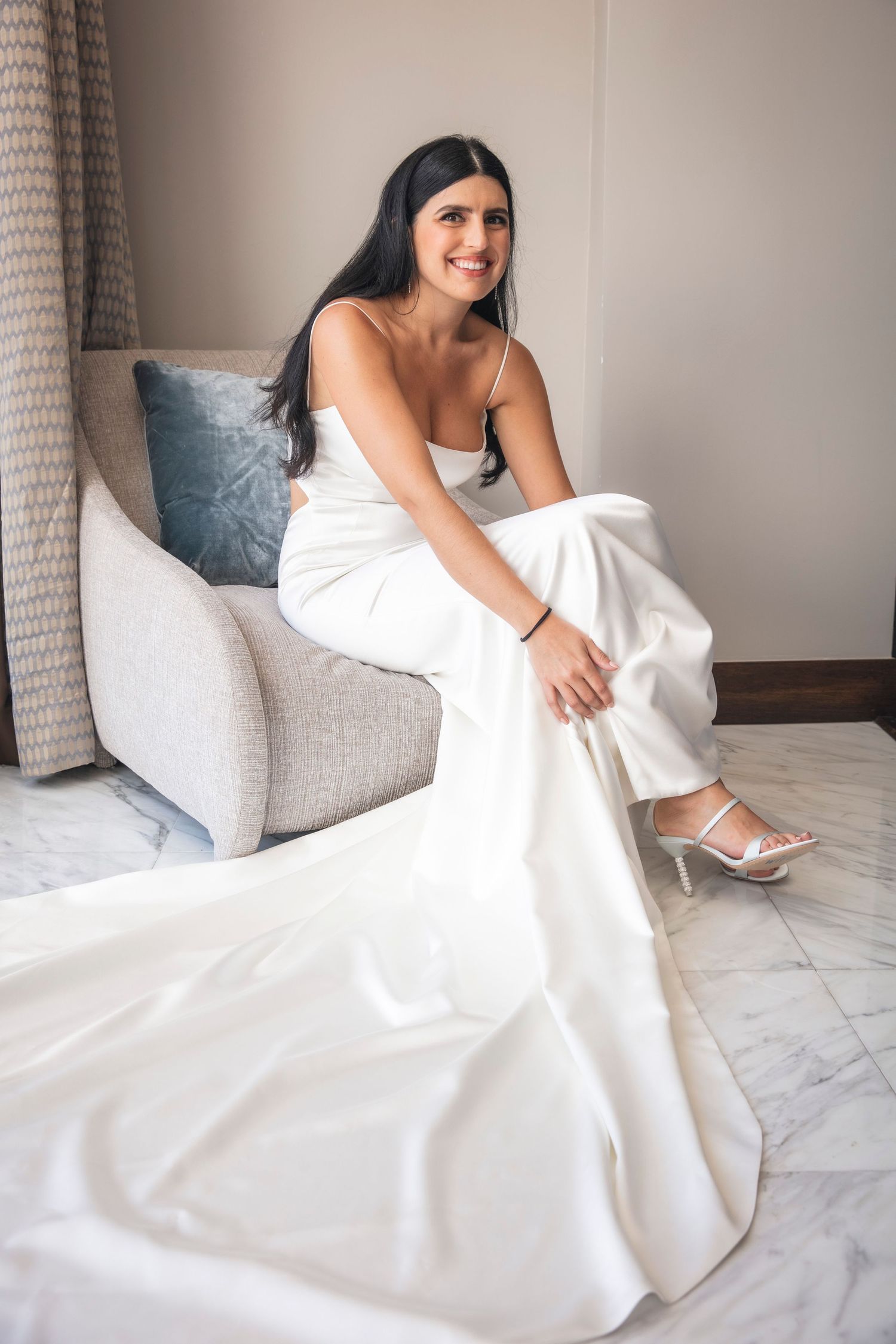 A bride in an elegant white wedding dress sits on a light gray couch smiling while showing off her white heels.