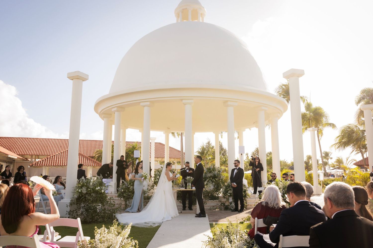 Wedding ceremony at an elegant beachfront gazebo with white domed architecture and palm trees at sunset.