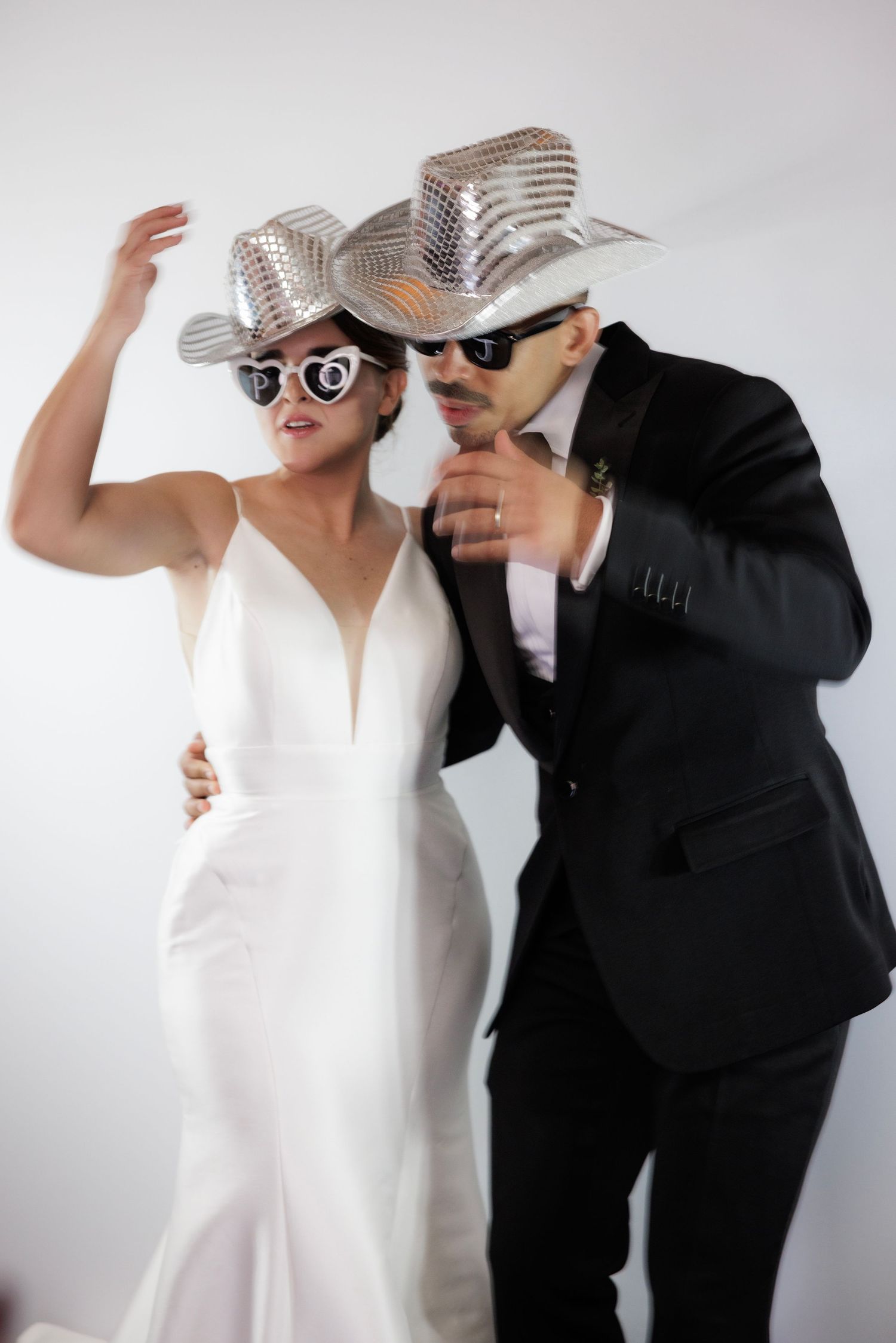 A couple poses playfully wearing matching striped fedoras and sunglasses in a wedding photoshoot series.