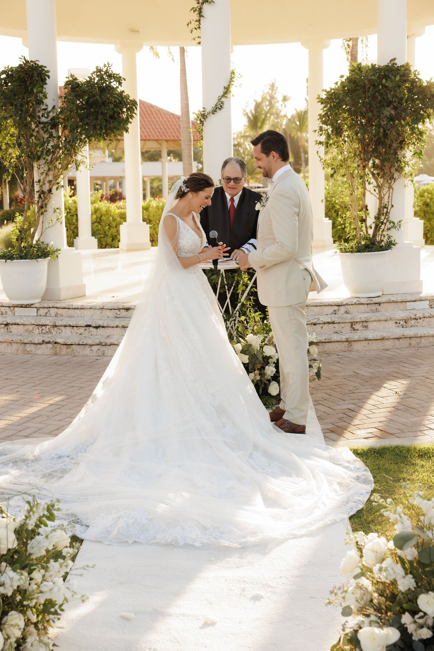 A wedding photo of a couple in a white gazebo with white pillars and greenery, bathed in warm sunlight.