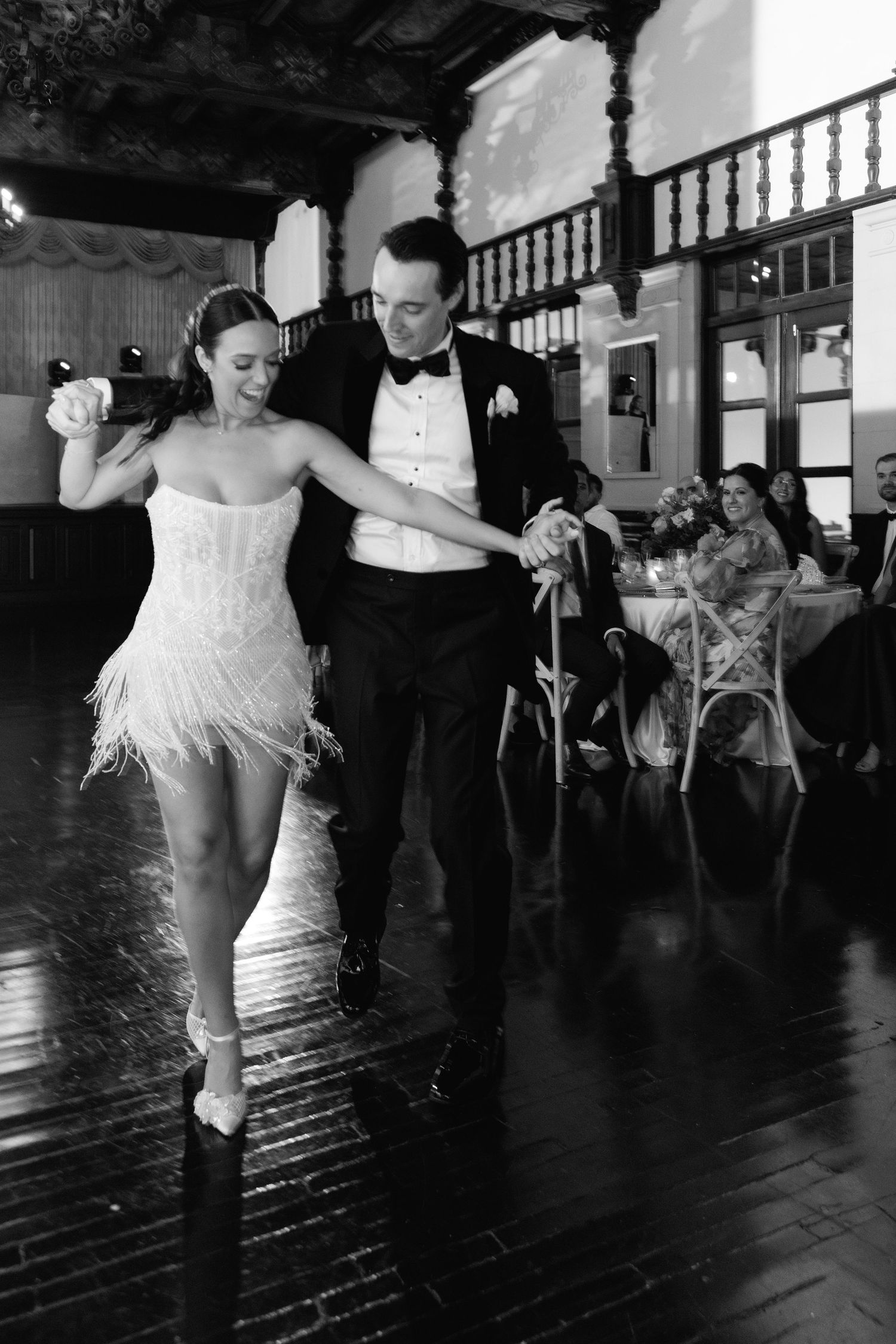 Dancers in formal attire perform dynamic moves on a reflective floor in a vintage ballroom setting.