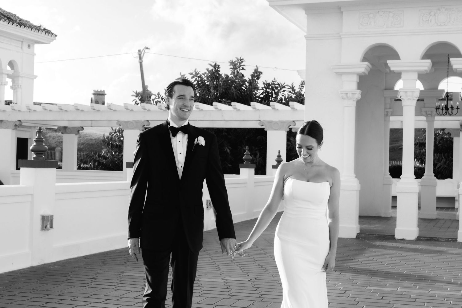 Bride in white dress stands with groom and wedding party under Mediterranean-style white archways in outdoor venue.