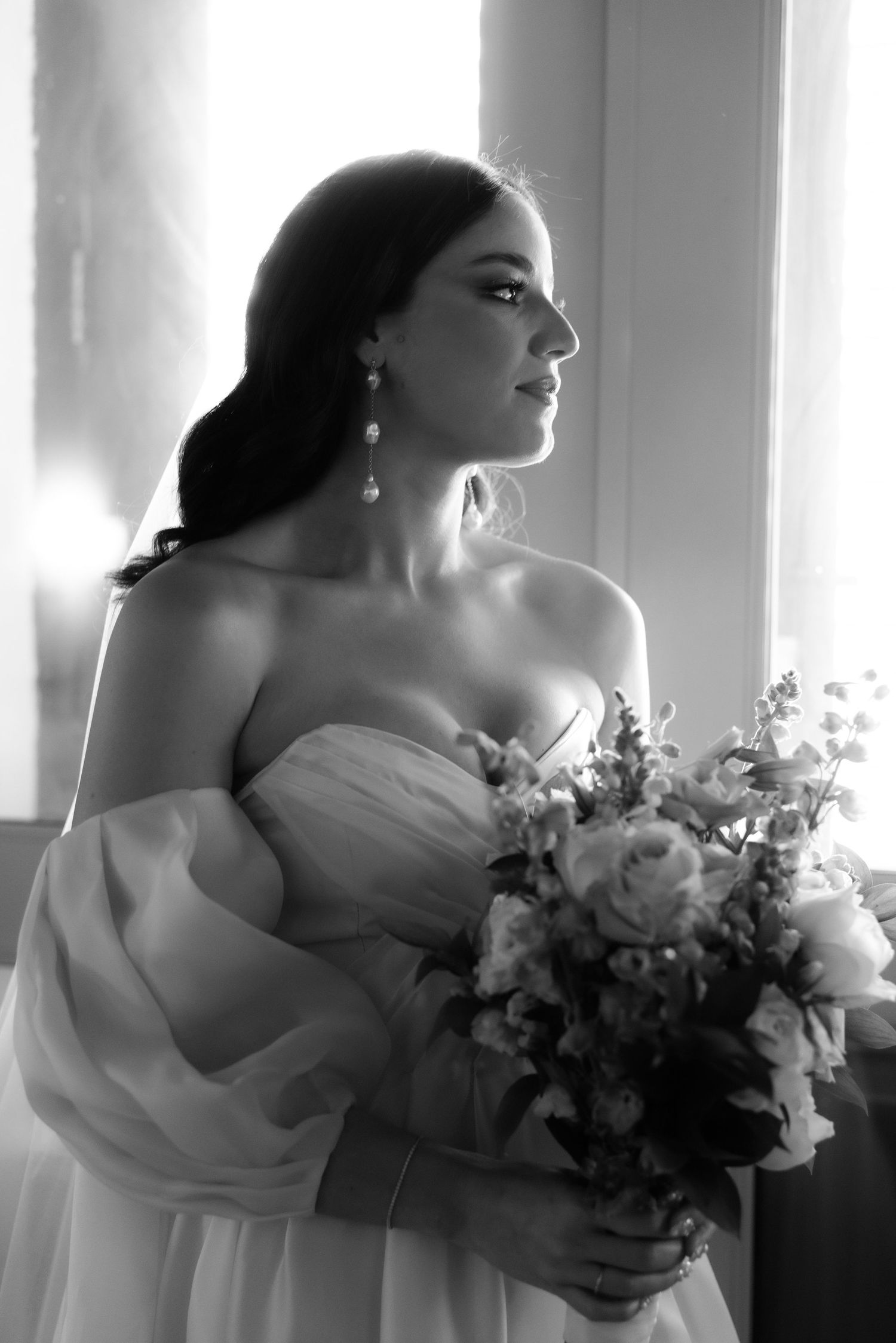Black and white portrait series of bride holding bouquet in elegant off-shoulder wedding dress near window.